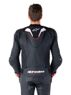 Alpinestars Missile V3 Ignition leren jas, achteraanzicht van een motorrijder, zwart leer met witte en rode accenten, voorzien van een slanke sport aero-hump met wit logo en 'alpinestars RACING AHEAD' branding op de onderrug, gecombineerd met bijpassende zwarte leren racebroek en handschoenen.