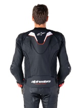 Rear view of a rider wearing the Alpinestars Missile V3 Ignition Leather Jacket, black leather with white and red accents, featuring a slim sport aero-hump with a white logo and 'alpinestars RACING AHEAD' branding on the lower back, paired with matching black leather racing pants and gloves.