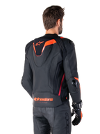 Alpinestars Missile V3 Ignition leren jas, heren race-sportjas, zwart met Red Fluo accenten, achteraanzicht met de slanke aerodynamische sporthump en Alpinestars-branding op de onderrug, gedragen met een zwarte leren broek en racehandschoenen, strak professioneel motorsportontwerp