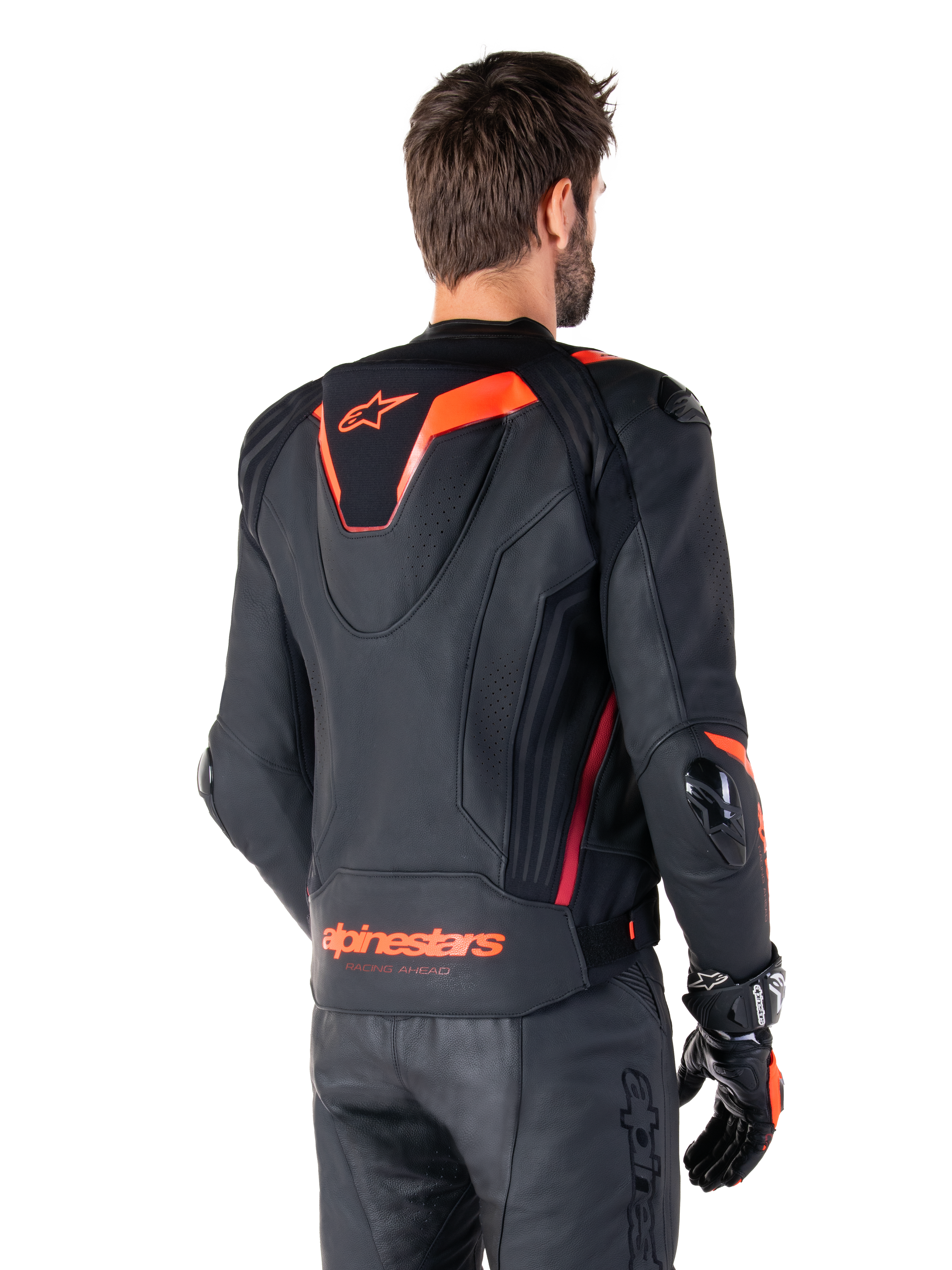Alpinestars Missile V3 Ignition leren jas, heren race-sportjas, zwart met Red Fluo accenten, achteraanzicht met de slanke aerodynamische sporthump en Alpinestars-branding op de onderrug, gedragen met een zwarte leren broek en racehandschoenen, strak professioneel motorsportontwerp