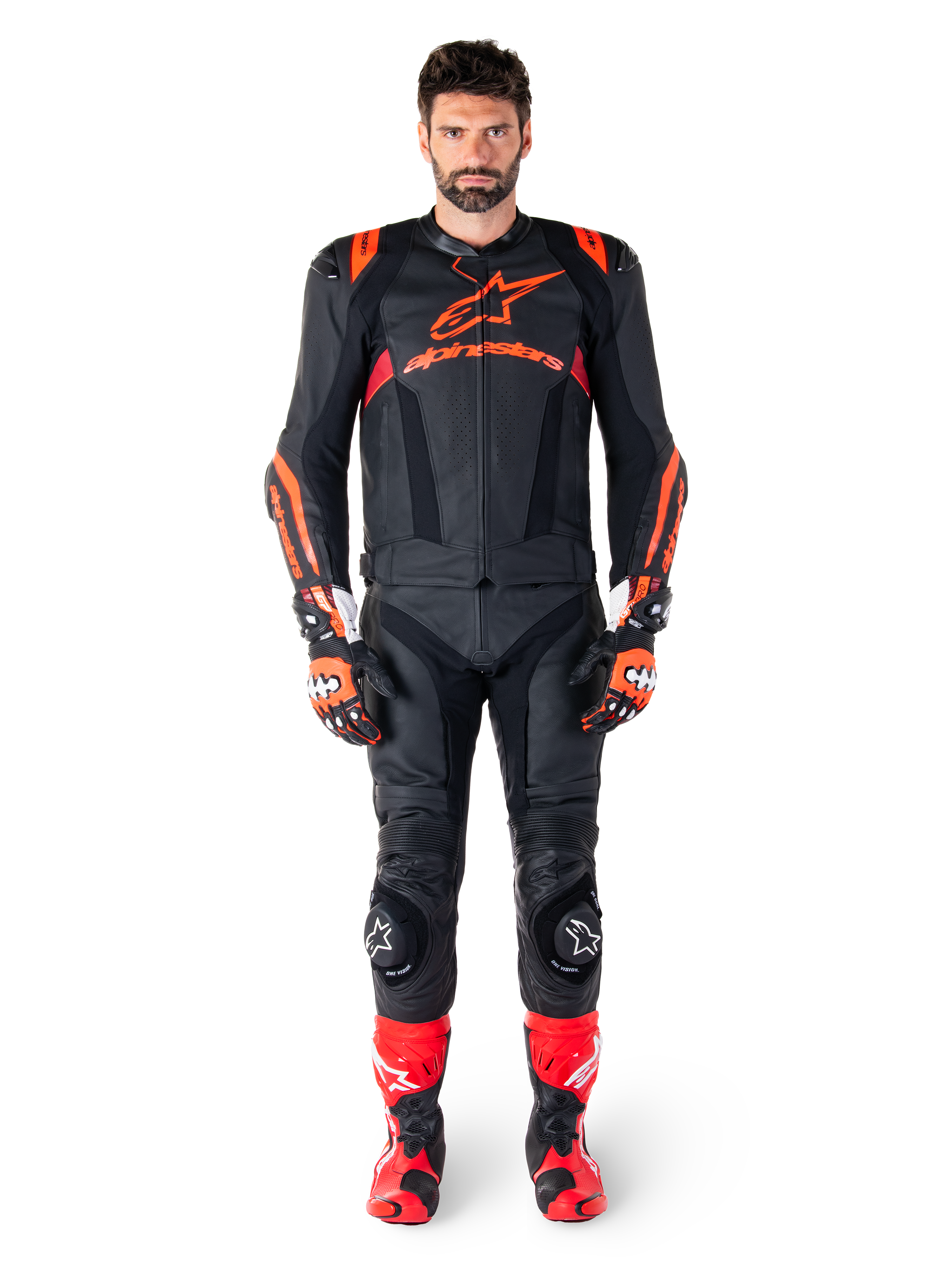 Mannelijk model draagt Alpinestars Missile V3 Ignition leren jas in Black Red Fluo Dark Red, complete motorrace-uitrusting inclusief bijpassende leren broek, racehandschoenen en laarzen, voorzien van opvallende Alpinestars-branding op de borst, naar voren gerichte pose, professionele sport-rijoutfit