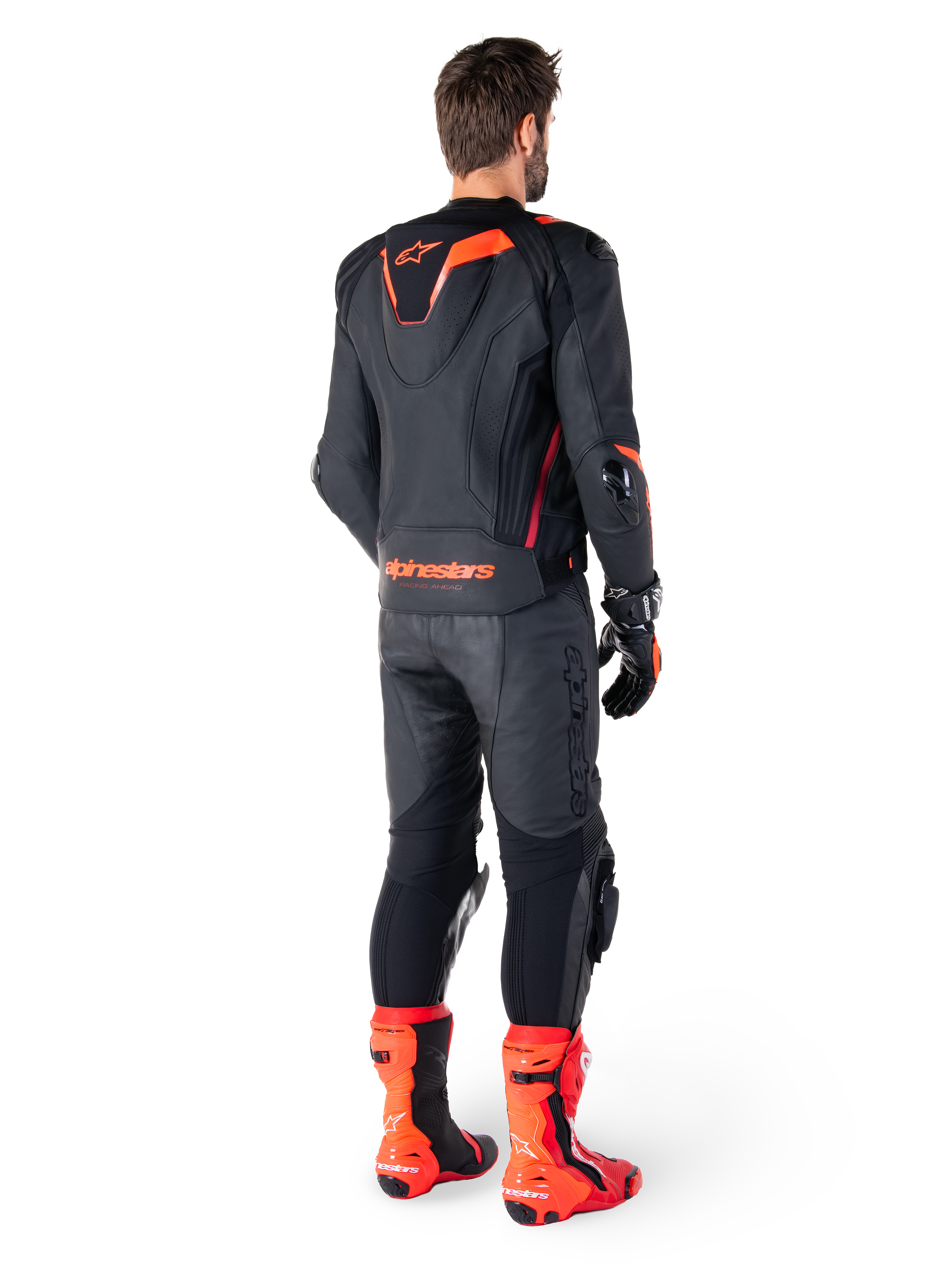 Alpinestars Missile V3 Ignition leren jas, race-sportmotorjas, zwart en fluo rood, achteraanzicht van een rijder die een volledige race-outfit draagt met bijpassende leren broek, handschoenen en rode laarzen, voorzien van een sport Aero-hump en opvallende merknaam op de onderrug.