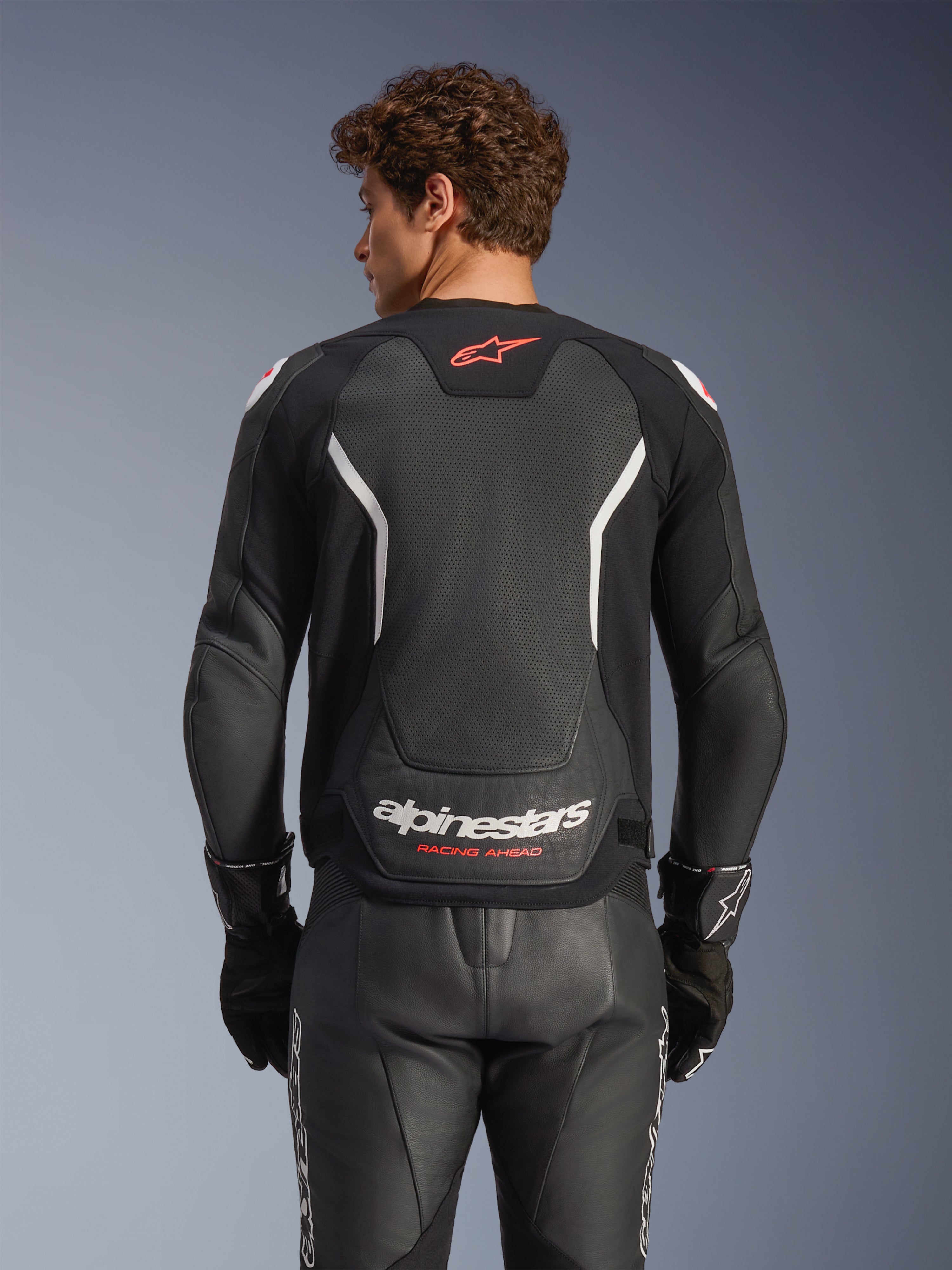 Alpinestars GP Force V2 Air Leren Jas, zwart met witte en rode accenten, achteraanzicht met geperforeerd leren rugpaneel, witte Alpinestars-branding en rode logo-details, gedragen met bijpassende zwarte leren racebroek en handschoenen.