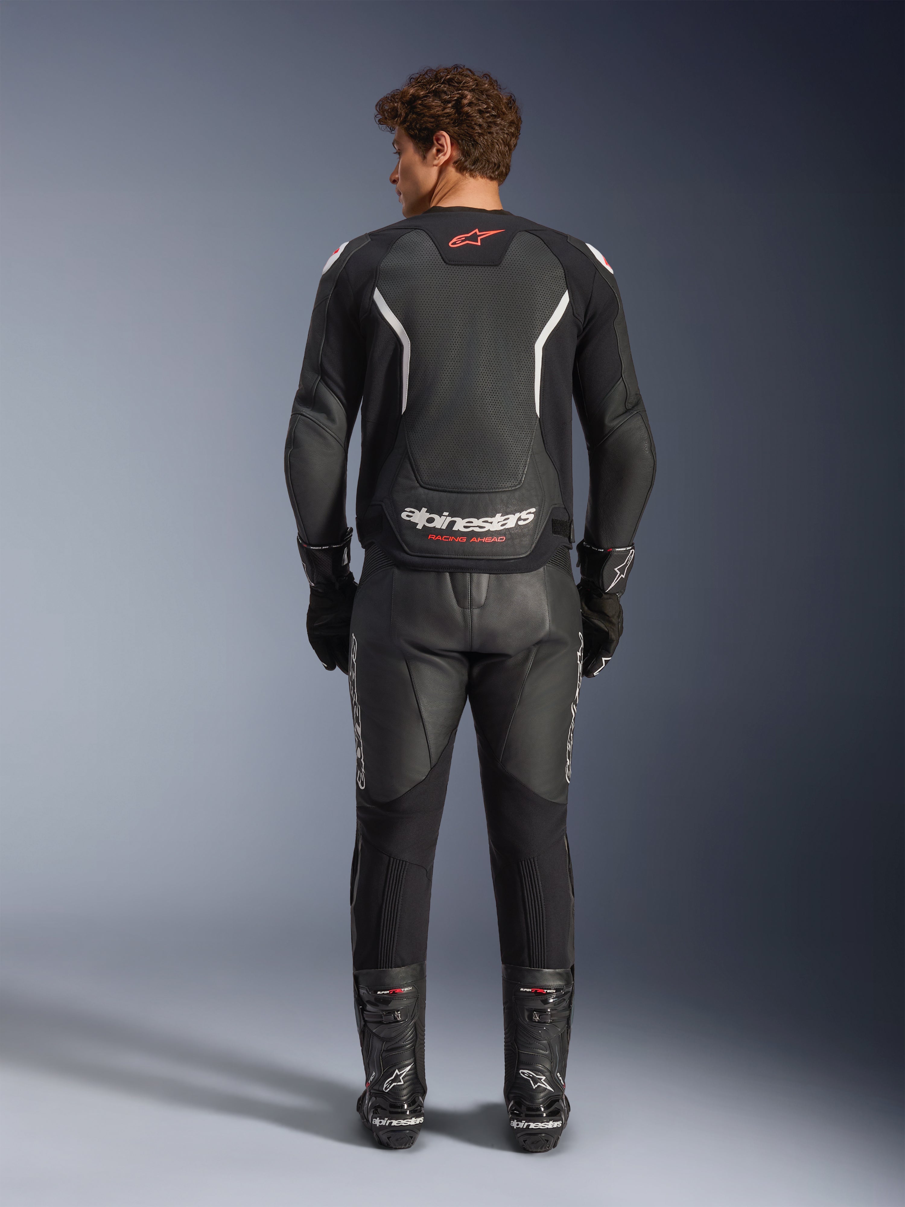 Alpinestars GP Force V2 Air Leren Jas in zwart en wit, achteraanzicht, voorzien van uitgebreide perforaties voor luchtstroom, witte schouderaccenten en opvallende branding, gedragen met een zwarte leren motorbroek en laarzen