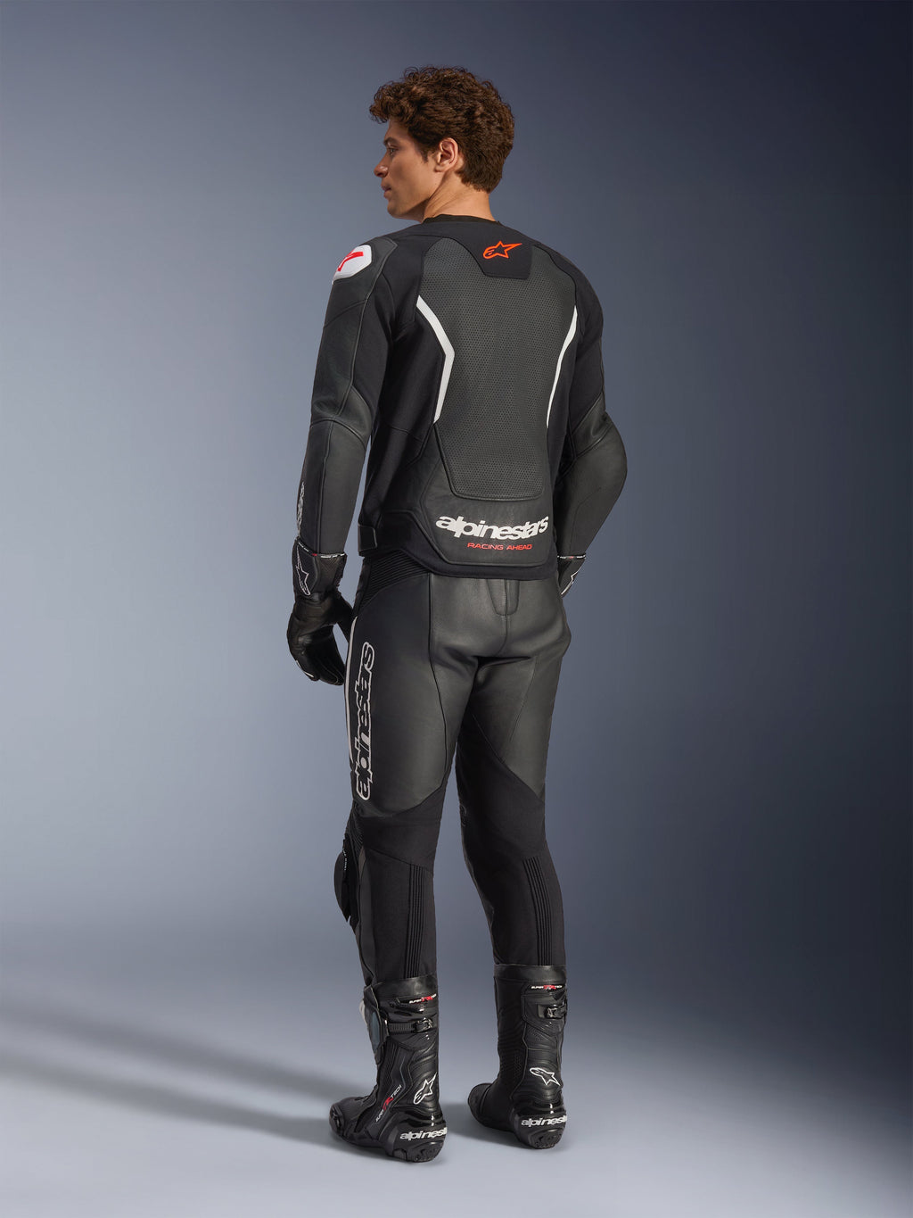 Alpinestars GP Force V2 Air leren jas, zwart en wit, achteraanzicht op model met geperforeerd rugpaneel voor ventilatie, witte branding en rood sterlogo, gedragen met bijpassende leren racebroek, handschoenen en laarzen