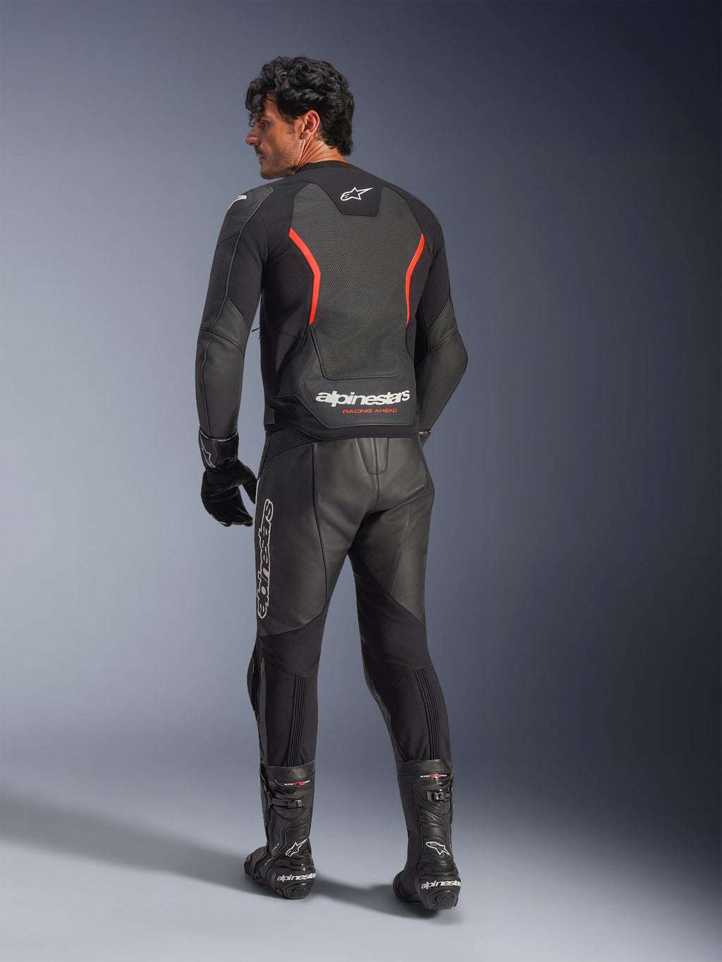 Rear view of a model wearing the Alpinestars GP Force V2 Air Leather Jacket in Black Red Fluo, featuring perforated leather panels and bold Alpinestars branding on the lower back, styled with matching black leather motorcycle pants and boots.
