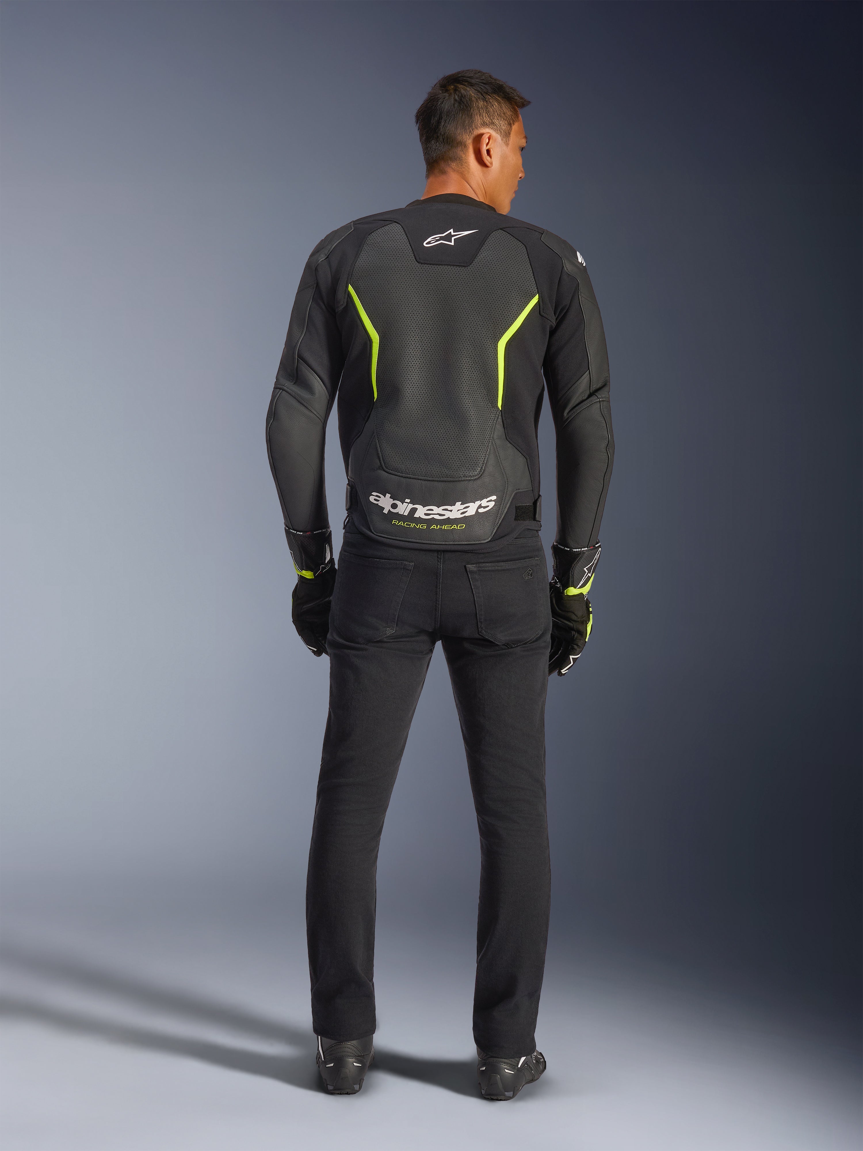 Alpinestars GP Force V2 Air Leren Jas, zwart en fluogeel, achteraanzicht van model dat de sportieve motorjas draagt, voorzien van grote geperforeerde delen, neongele accenten en witte 'alpinestars' branding op de onderrug, gecombineerd met zwarte motoruitrusting