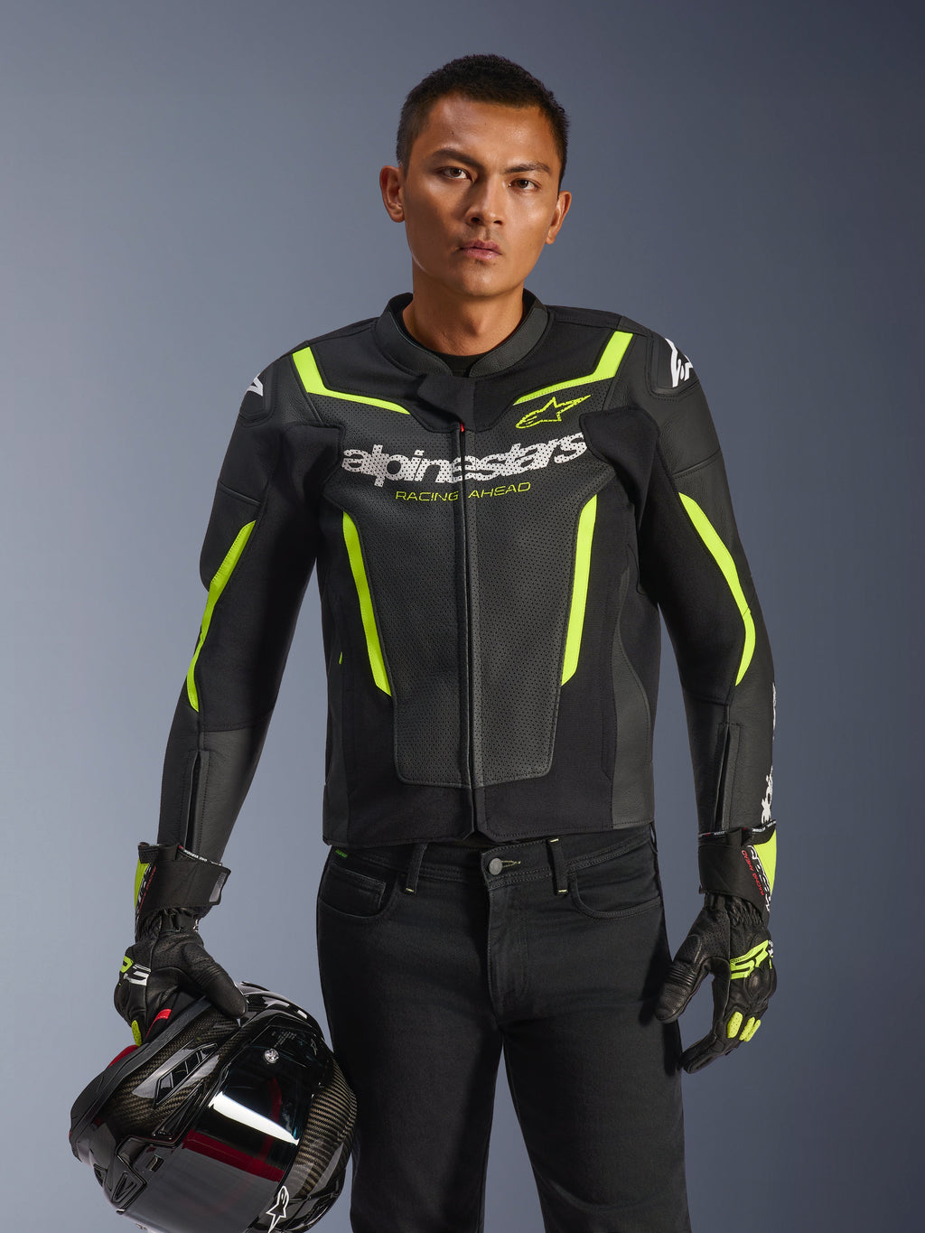 Mannelijke motorrijder draagt de Alpinestars GP Force V2 Air Leren Jas in de kleur zwart en neongeel, houdt een zwarte motorhelm vast, voorzien van geperforeerd leer met witte Alpinestars-branding en fluogele accenten, gecombineerd met bijpassende racehandschoenen en een zwarte broek