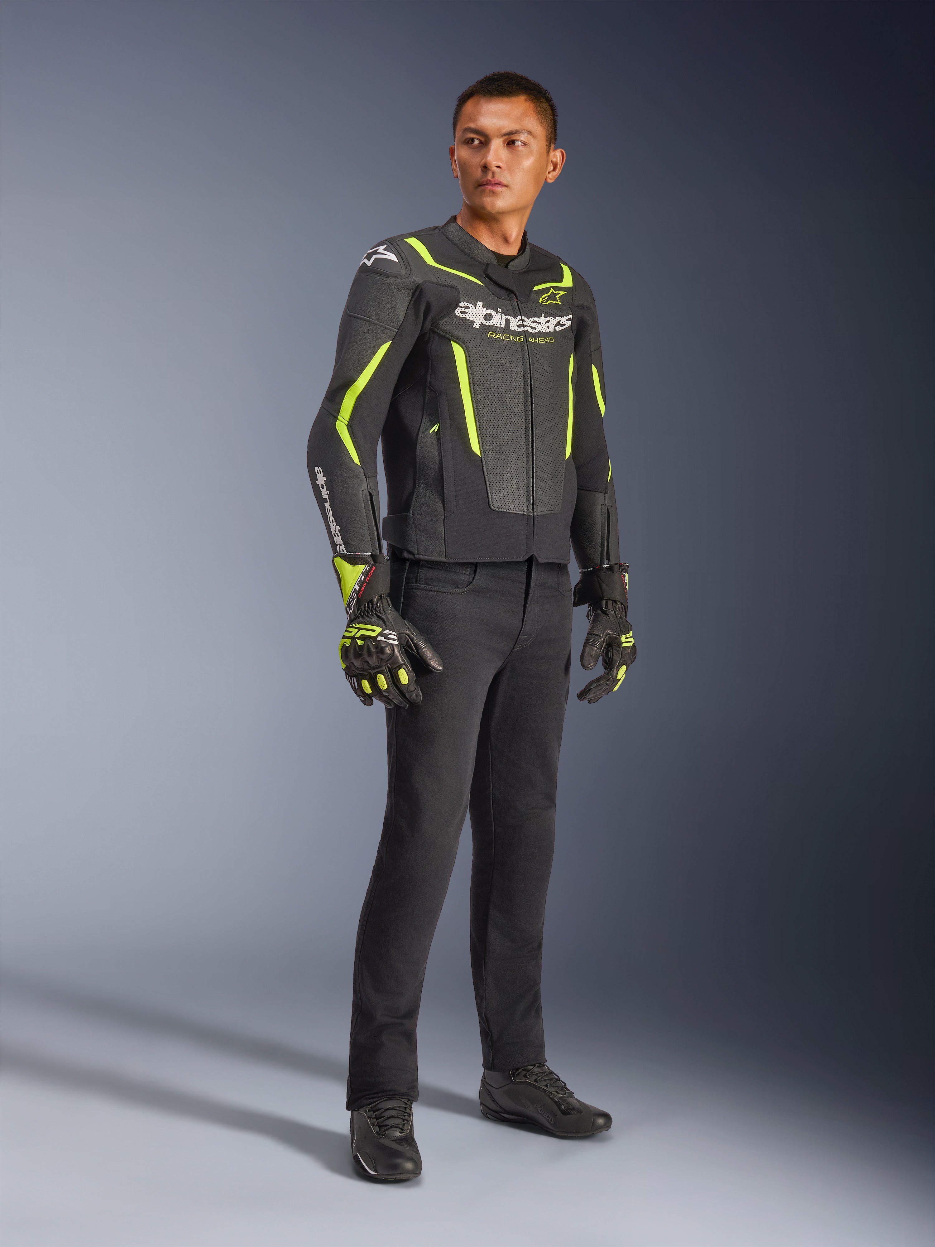 Mannelijk model draagt Alpinestars GP Force V2 Air Leren Jas in zwart en fluogeel, gecombineerd met zwarte motorbroek, bijpassende handschoenen en zwarte schoenen, volledige studio-opname van sportieve motoruitrusting met geperforeerd leer en opvallende branding