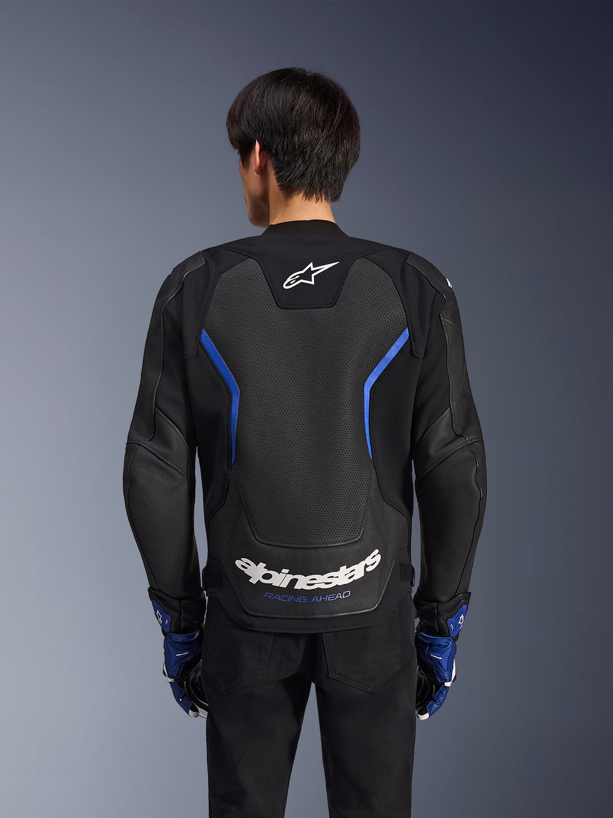 Alpinestars GP Force V2 Airflow leren jas, zwart metallic blauw, achteraanzicht, voorzien van een groot geperforeerd rugpaneel voor ventilatie, blauwe accenten en witte Alpinestars-branding, gedragen met bijpassende blauwe handschoenen en een zwarte broek voor sportief rijden.