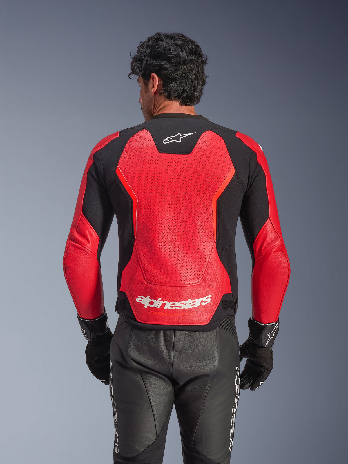 Alpinestars GP Force V2 Air Leren Jas in rood en zwart, achteraanzicht met het geperforeerde leren chassis voor luchtstroom, witte Alpinestars-branding op de onderrug en geïntegreerde stretchpanelen, gedragen met een zwarte leren motorbroek en handschoenen.