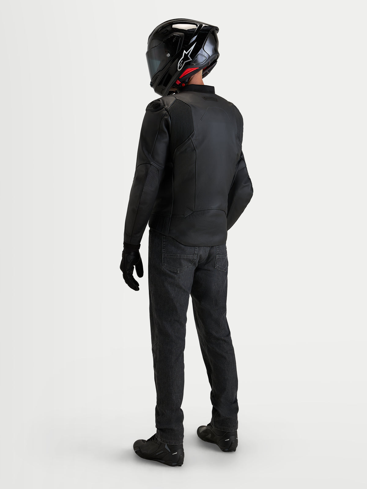Alpinestars Caliber V 2 leren jas, zwarte heren motorjas, achteraanzicht van een motorrijder in volledige uitrusting met helm en zwarte denim jeans