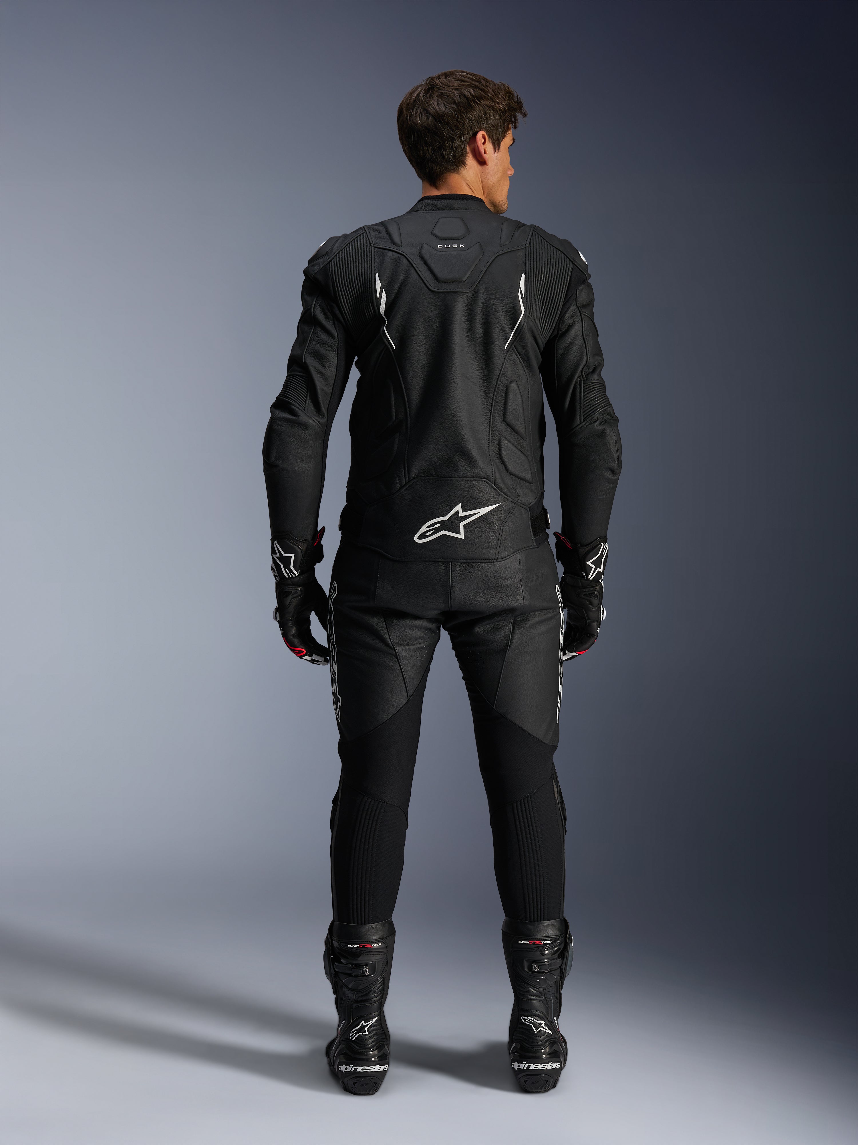 Alpinestars motorrijder van achteren gezien, Dusk Leather Jacket en Missile V3 Leather Pants, volledig zwart leren pak met witte accenten, jas met 'Dusk' branding en Alpinestars logo op de rug, gecombineerd met zwarte racelaarzen en handschoenen, gecoördineerde motorsport outfit voor wegraces