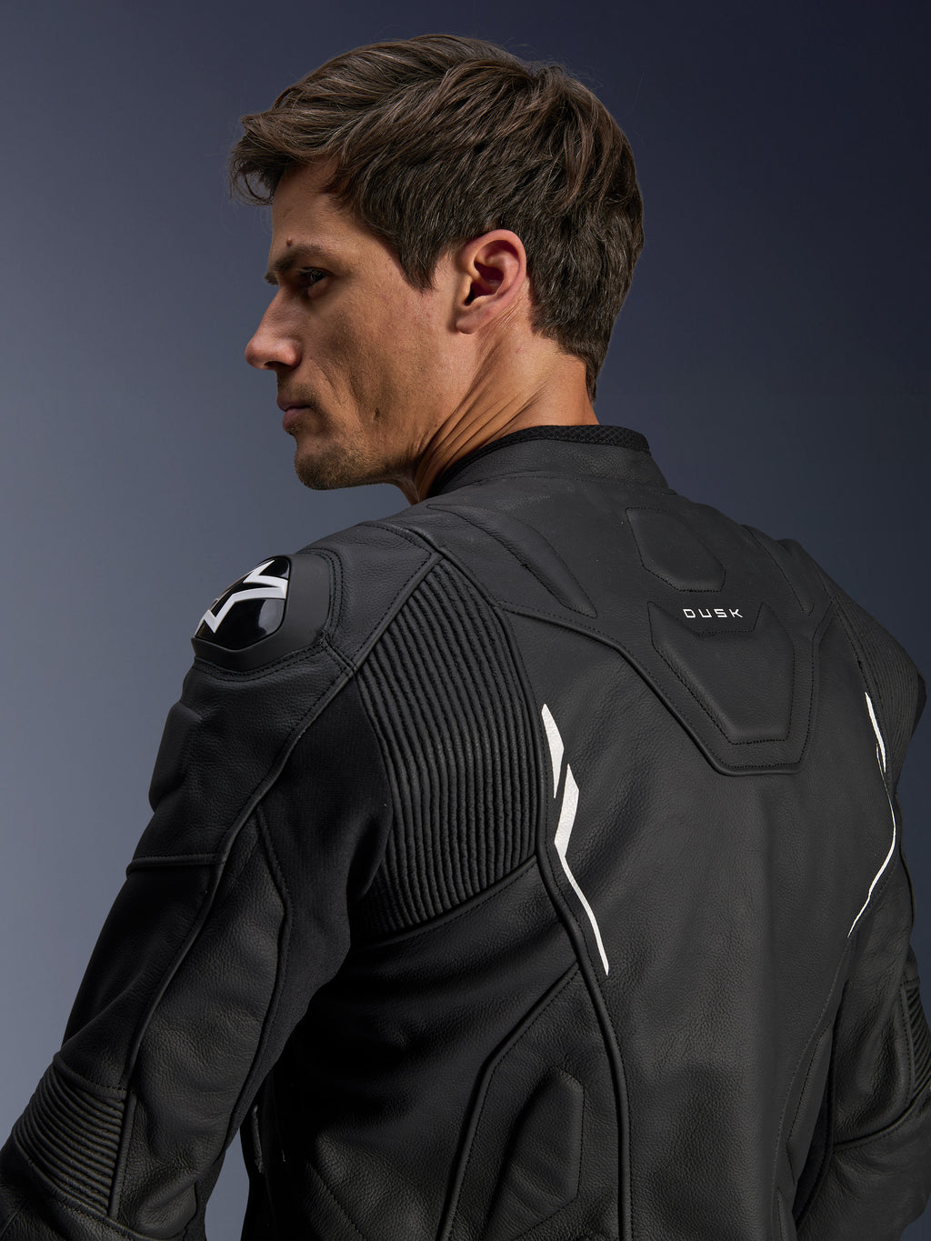 Alpinestars Dusk Leren Jas, zwart met witte accenten, achteraanzicht op model, voorzien van DFS schouderprotectoren met Alpinestars-logo, accordeon lederen stretchpanelen voor bewegingsvrijheid en 'DUSK' branding op de bovenrug, premium runderleren motorjas voor weg- en sportgebruik