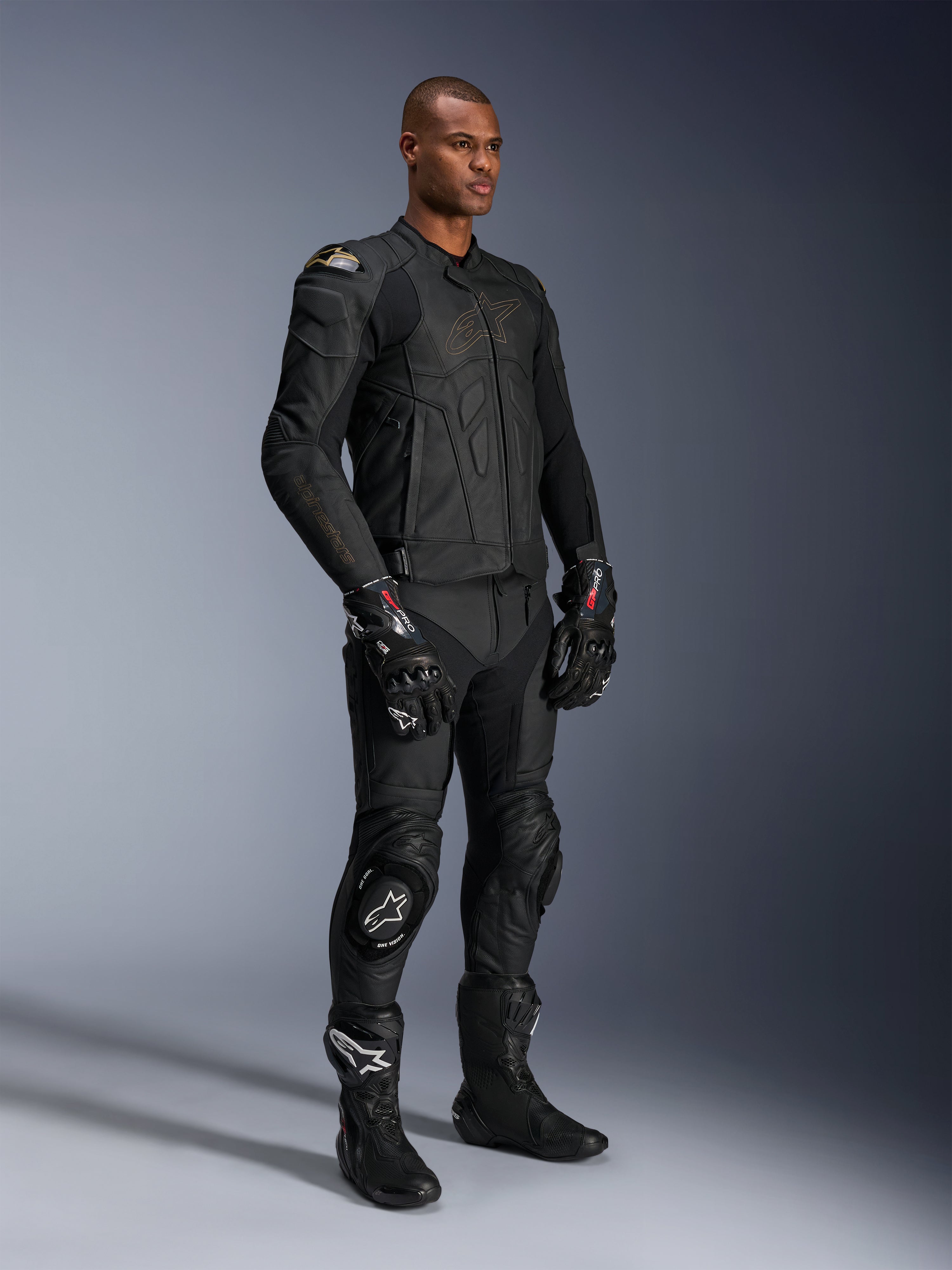Mannelijk model draagt de Alpinestars Dusk leren jas in Black Laser, gecombineerd met bijpassende zwarte leren racebroek, GP Pro handschoenen en performance circuitlaarzen. Volledige motorrace-uitrusting met externe DFS schouderbescherming en een strakke atletische pasvorm voor sportief rijden en op de weg.