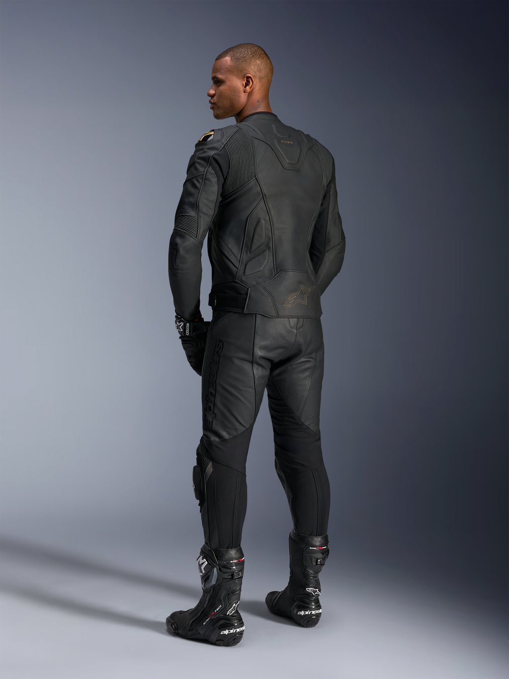 Alpinestars Dusk Leren Jas, motorjas, Black Laser, achteraanzicht op een model in volledige leren race-uitrusting, 3D multi-panel constructie met laser-geëtste branding, hoogwaardig runderleer voor sportief en straatgebruik.