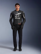 Mannelijk model draagt Alpinestars GP Plus V4 leren jas, zwart en wit, met opvallende witte Astars-branding op de borst en witte accenten op de schouders, gecombineerd met zwarte motorhandschoenen en broek, sportieve motoruitrusting