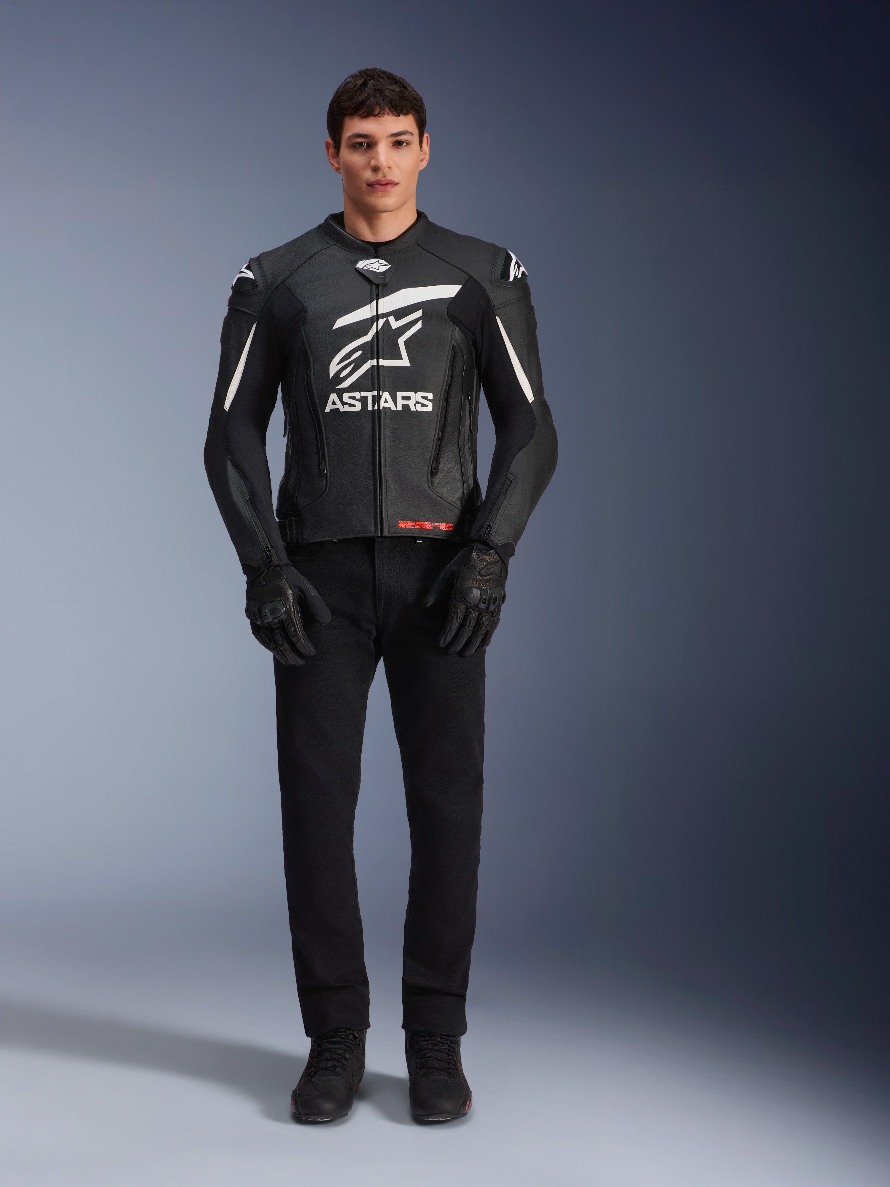 Mannelijk model draagt Alpinestars GP Plus V4 leren jas, zwart en wit, met opvallende witte Astars-branding op de borst en witte accenten op de schouders, gecombineerd met zwarte motorhandschoenen en broek, sportieve motoruitrusting
