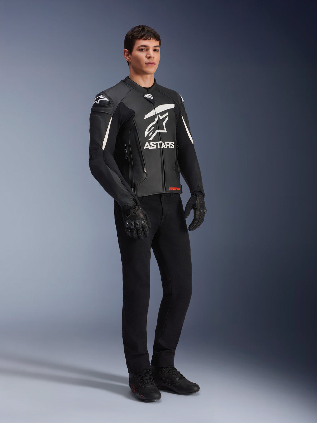 Alpinestars GP Plus V4 Leren Jas, sport motorjas, zwart en wit, gedragen door een mannelijk model met zwarte motorbroek en handschoenen, groot wit ASTARS-logo op de borst, externe DFS-schouder sliders, professioneel race-afgeleid ontwerp