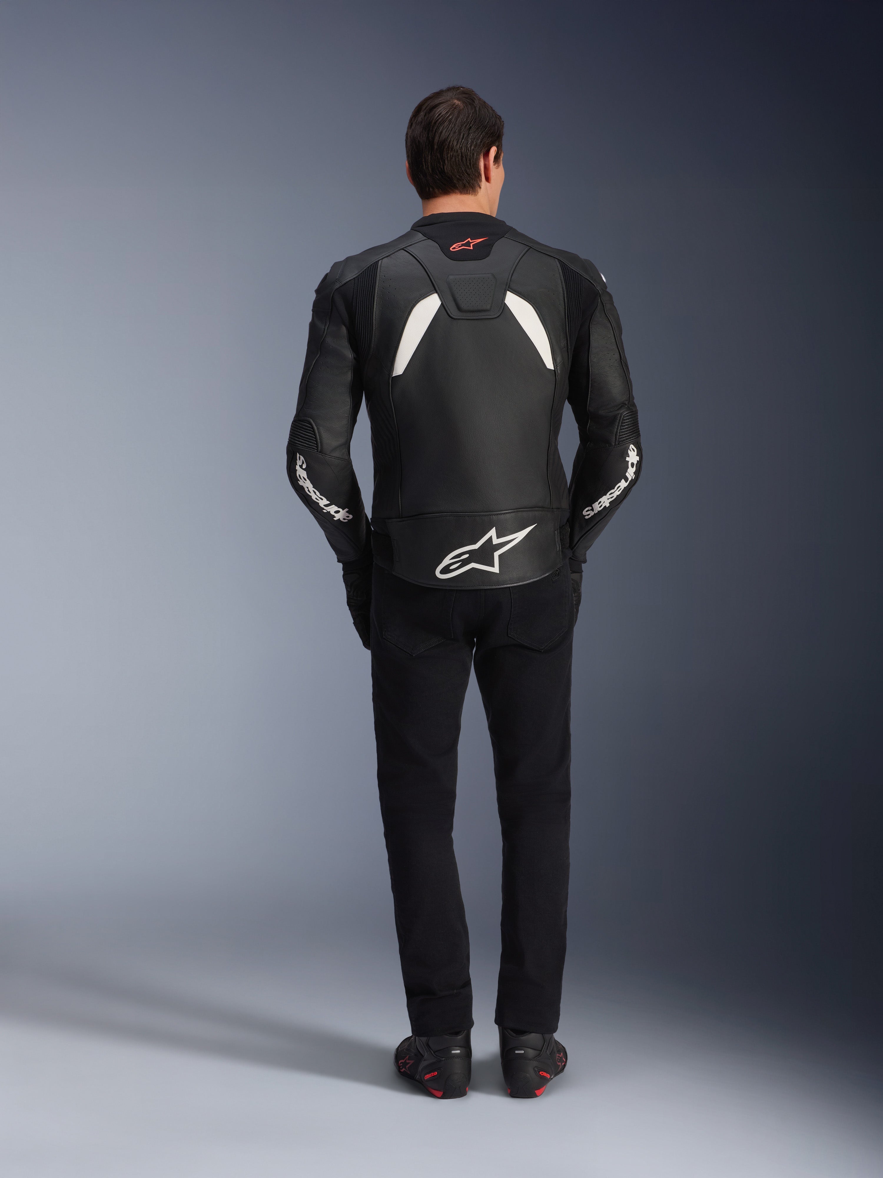 Alpinestars GP Plus V4 leren jas in zwart en wit, model van achteren gezien, gecombineerd met een zwarte broek en motorlaarzen, voorzien van witte panelen op de schouders en Alpinestars-logo's op de mouwen en onderrug.