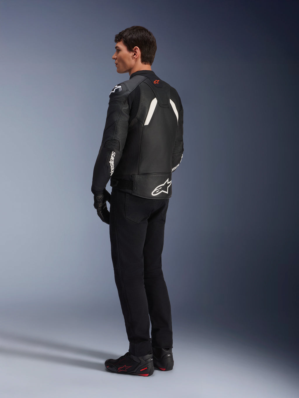 Alpinestars GP Plus V4 Leren Jas, sportief leren jack, zwart en wit, gedragen door een model in zijaanzicht van achteren, met witte Alpinestars-logo's op de onderrug en mouw, gecombineerd met een zwarte motorbroek en technische laarzen voor een sportieve look.