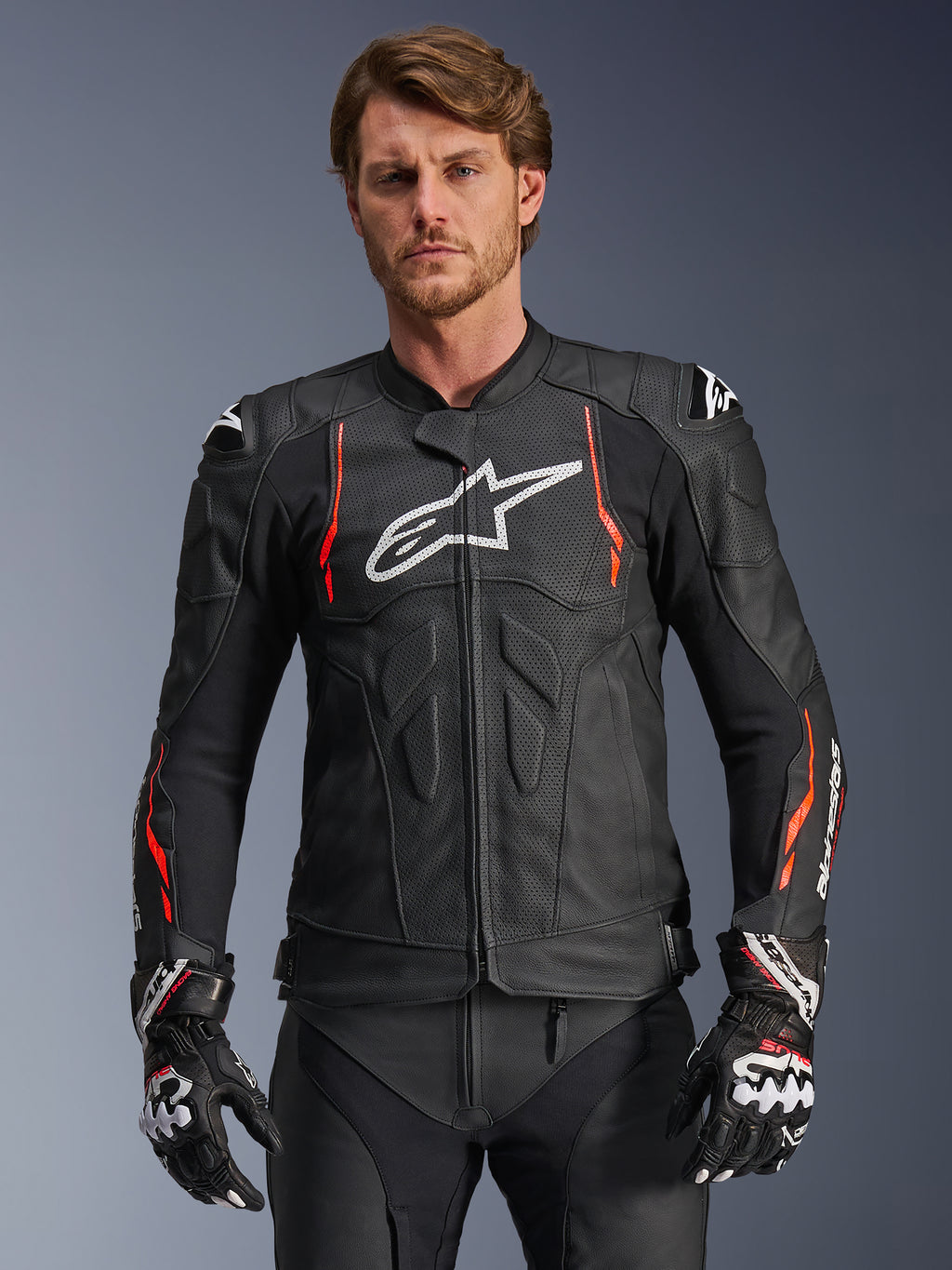 Alpinestars Dusk Airflow leren motorjas, zwart met fluorescerend rode en witte accenten, lederen motorracejas voorzien van uitgebreide perforaties voor ventilatie, wit logo op de borst en externe schoudersliders, gedragen door een model met bijpassende racehandschoenen