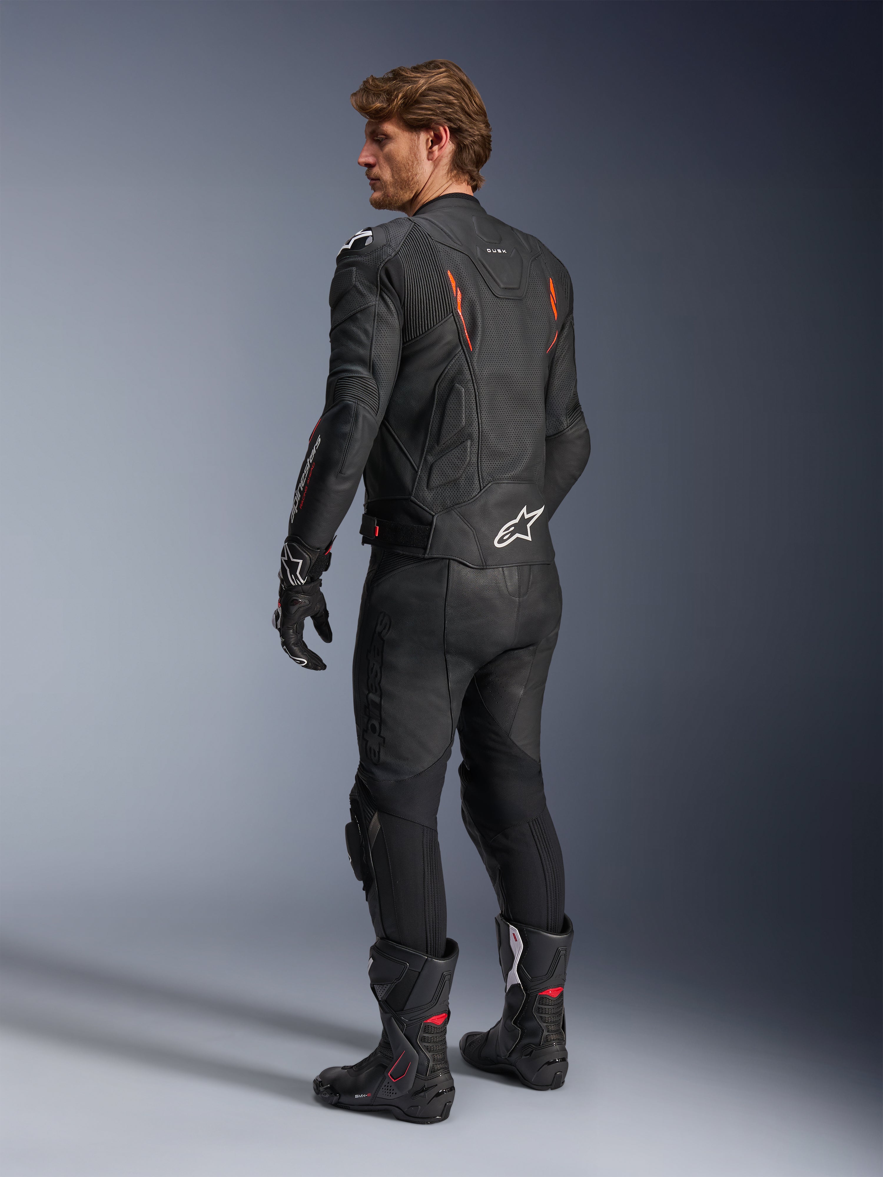 Alpinestars Dusk Airflow Leren Jas, zwart met Red Fluo accenten, achteraanzicht van een rijder in volledige leren motoruitrusting inclusief bijpassende zwarte broek, handschoenen en racelaarzen, voorzien van geperforeerd leer en witte Alpinestars-logo's.