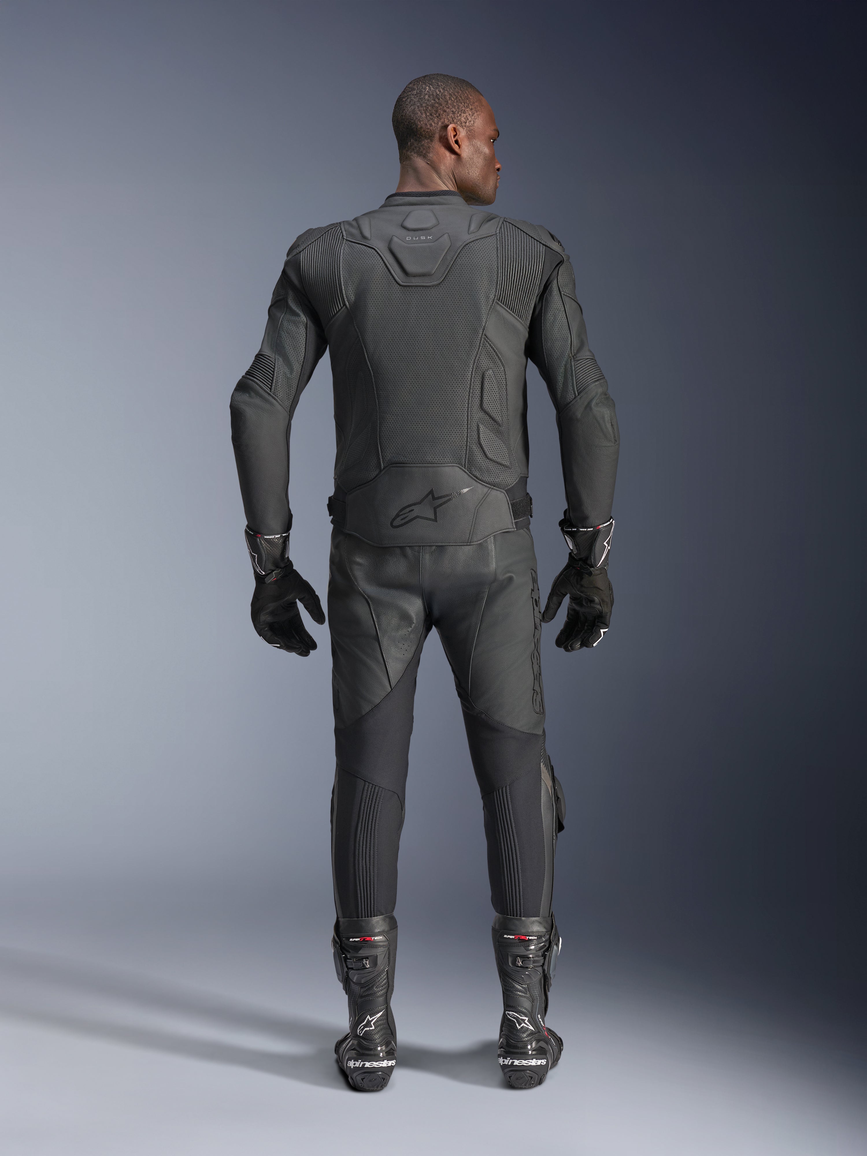 Mannelijk model van achteren getoond, draagt de Alpinestars Dusk Airflow leren jas in het zwart, gecombineerd met bijpassende zwarte leren racebroek, handschoenen en laarzen, voorzien van uitgebreide perforaties voor ventilatie en het Alpinestars-logo op de onderrug.