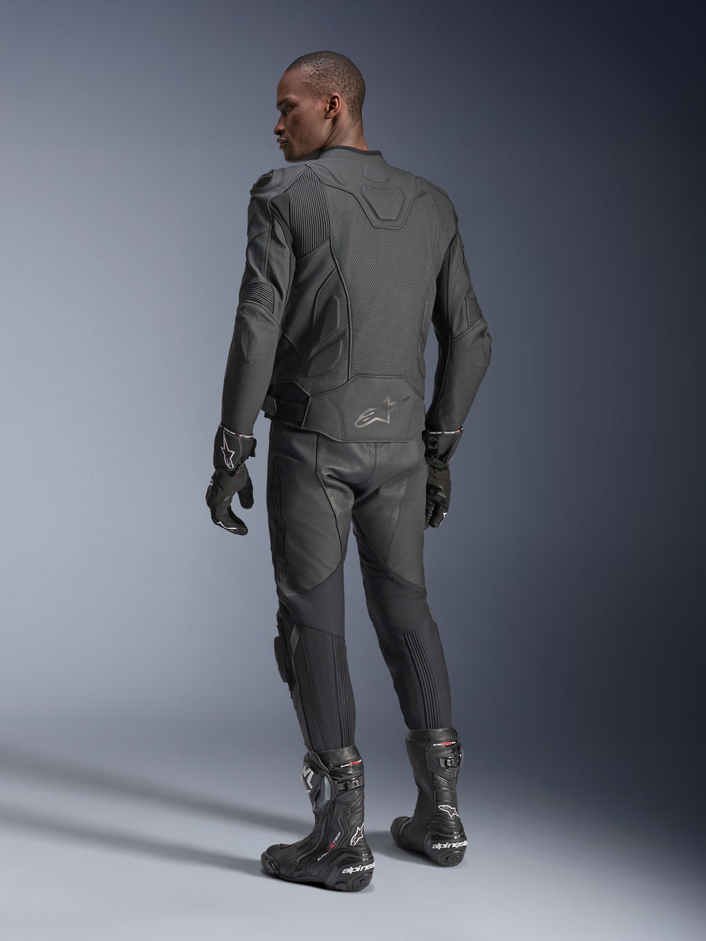 Mannelijke rijder van achteren die de Alpinestars Dusk Airflow Leren Jas in Black Black draagt, gecombineerd met een bijpassende leren broek, handschoenen en laarzen, wat de uitgebreide perforaties op de rug en de sportieve pasvorm benadrukt.