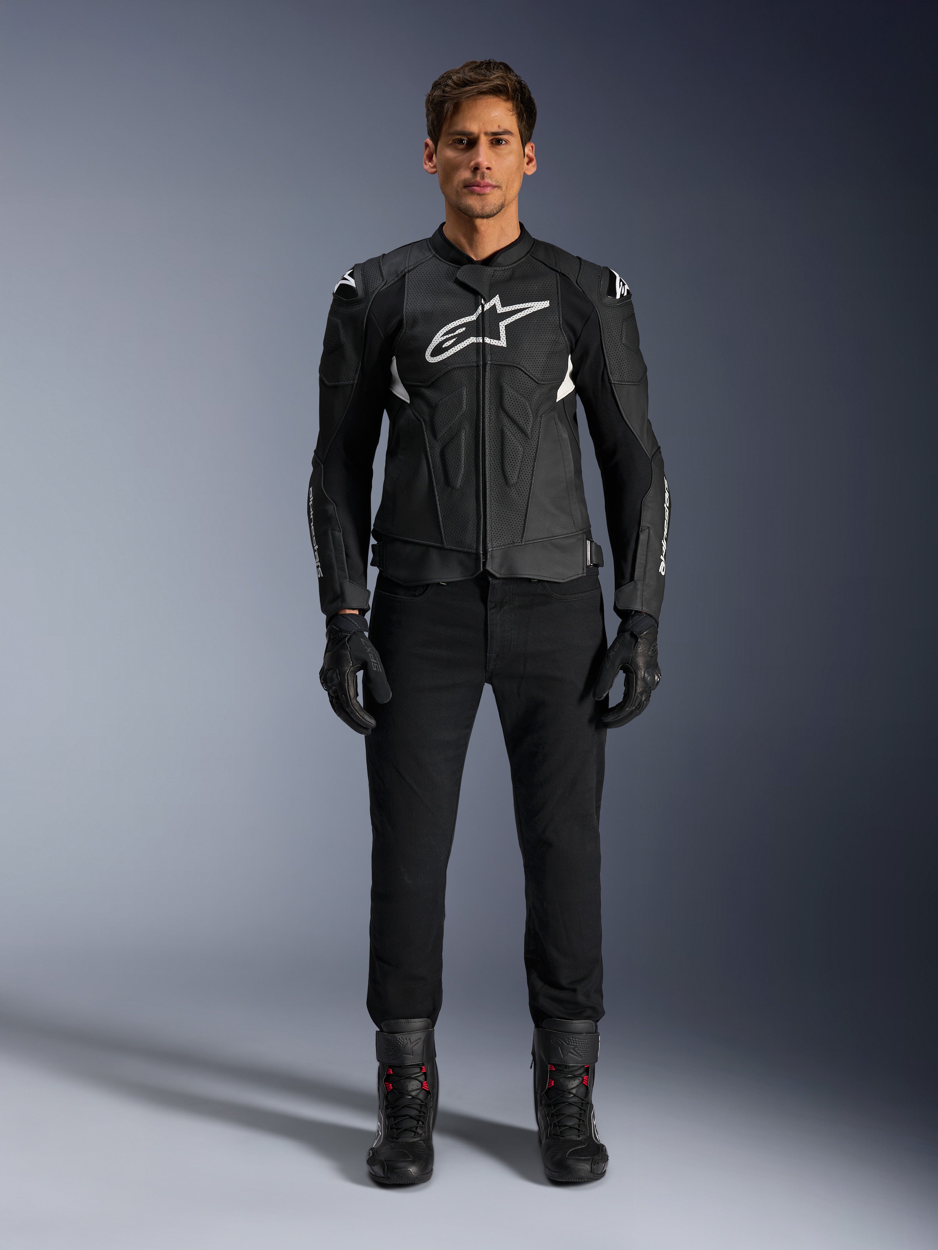 Alpinestars Dusk Airflow Leren Jas, zwart en wit, leren motorjas met geperforeerde panelen voor luchtstroom, wit Astars-logo op de borst, gedragen door een mannelijke rijder met zwarte broek, handschoenen en motorlaarzen, professionele studio-opname voor sportieve motoruitrusting.