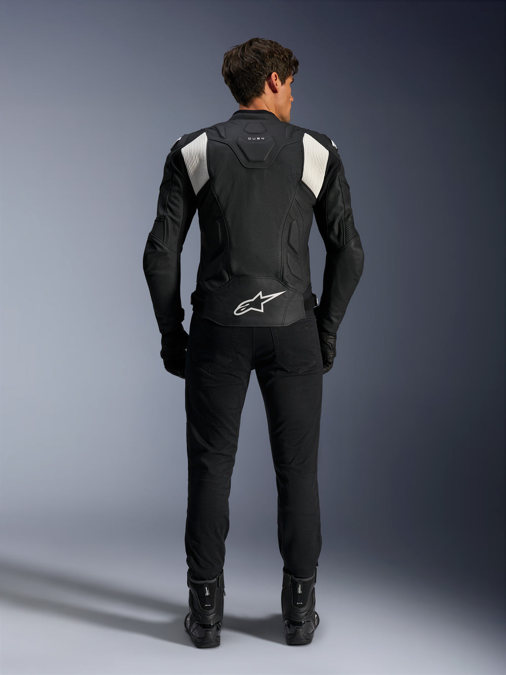 Alpinestars Dusk Airflow Leren Jas, Zwart en Wit, achteraanzicht van een rijder die een zwarte broek en laarzen draagt, voorzien van geperforeerd leer voor ventilatie, witte accordeon stretchpanelen op de schouders en witte branding op de onderrug