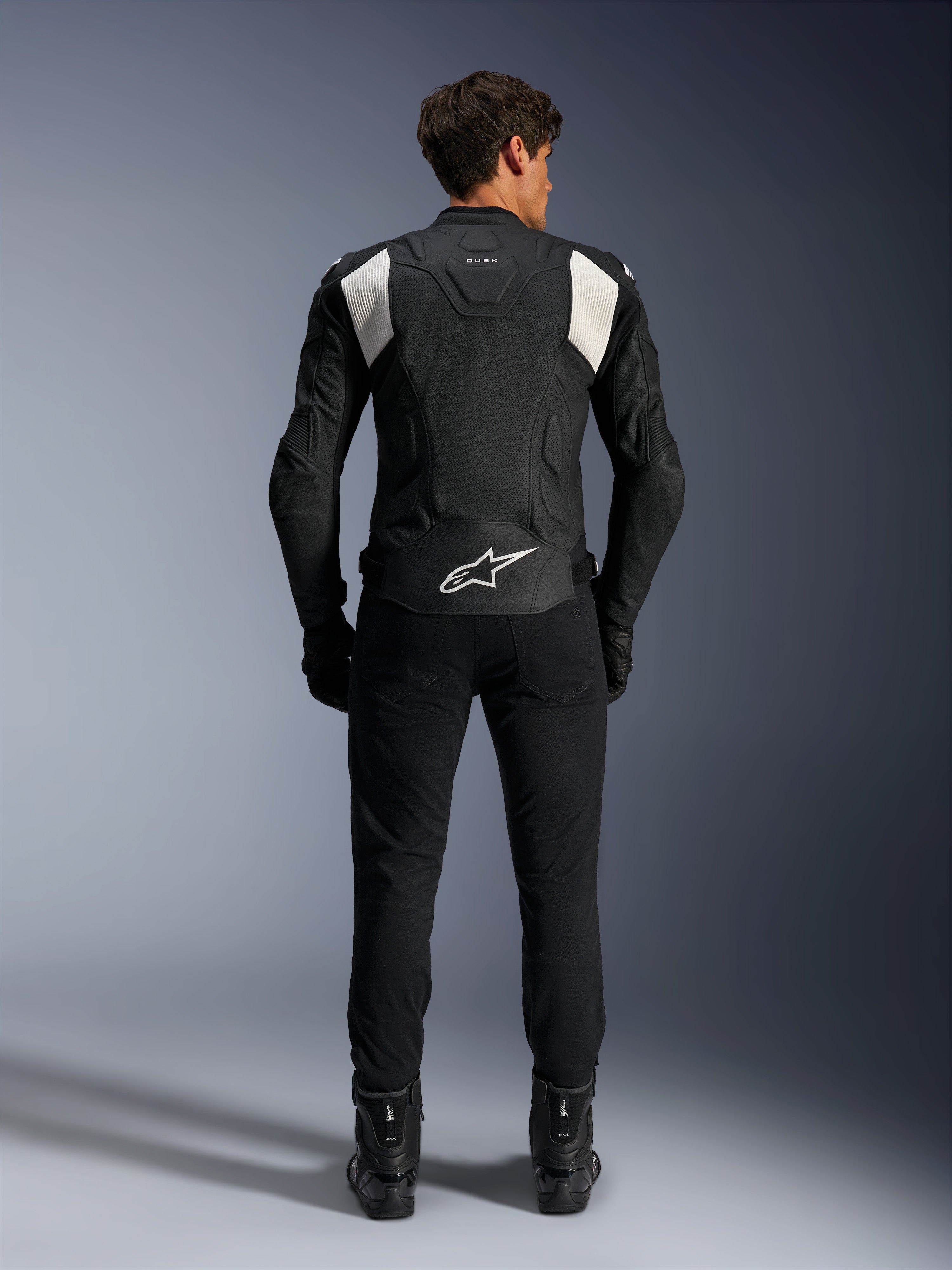 Alpinestars Dusk Airflow Leren Jas, Zwart en Wit, achteraanzicht van een rijder die een zwarte broek en laarzen draagt, voorzien van geperforeerd leer voor ventilatie, witte accordeon stretchpanelen op de schouders en witte branding op de onderrug
