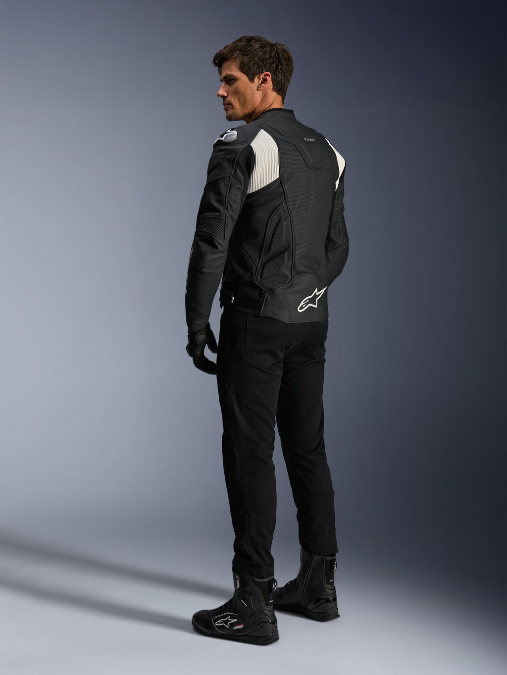 Alpinestars Dusk Airflow Leren Jas, Zwart en Wit, achteraanzicht van model die de geperforeerde leren jas draagt met bijpassende zwarte broek en laarzen, voorzien van witte accordeon stretchpanelen en een opvallend logo op de onderrug, sportieve motorrijuitrusting
