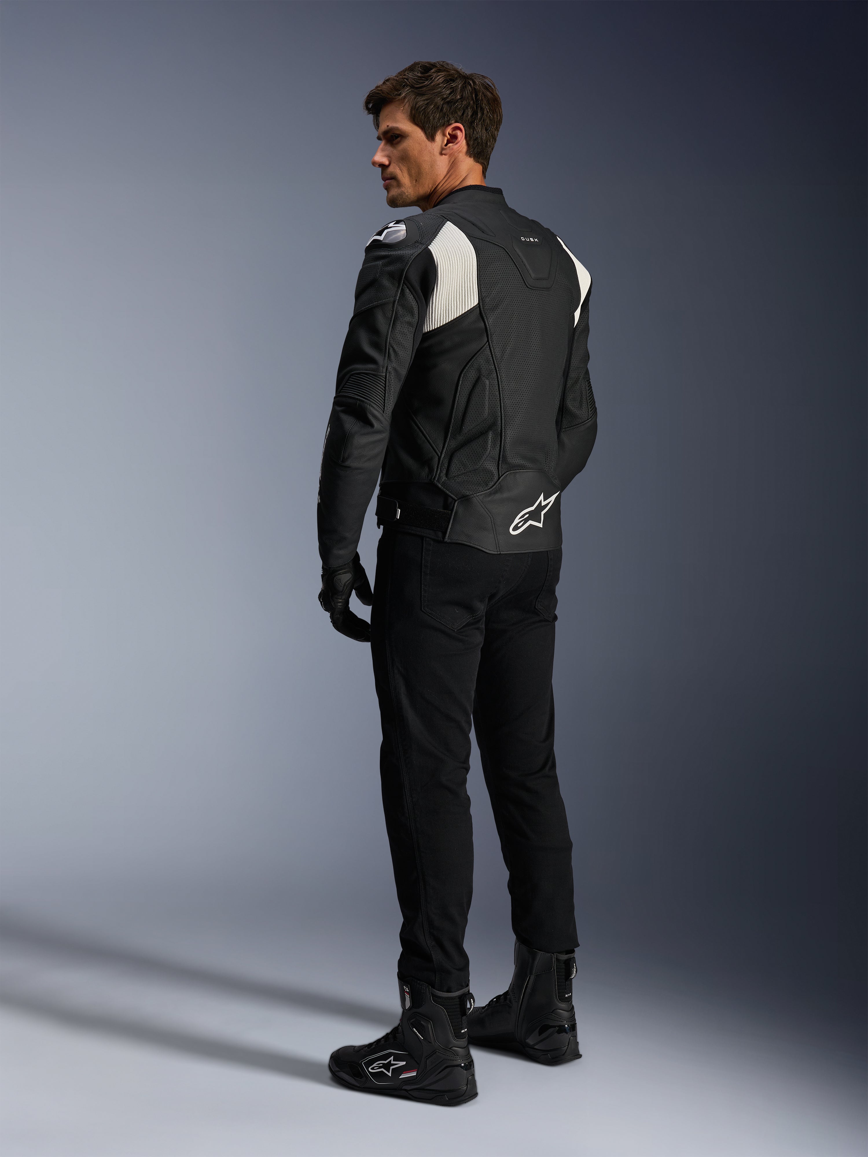 Alpinestars Dusk Airflow Leren Jas, Zwart en Wit, achteraanzicht van model die de geperforeerde leren jas draagt met bijpassende zwarte broek en laarzen, voorzien van witte accordeon stretchpanelen en een opvallend logo op de onderrug, sportieve motorrijuitrusting
