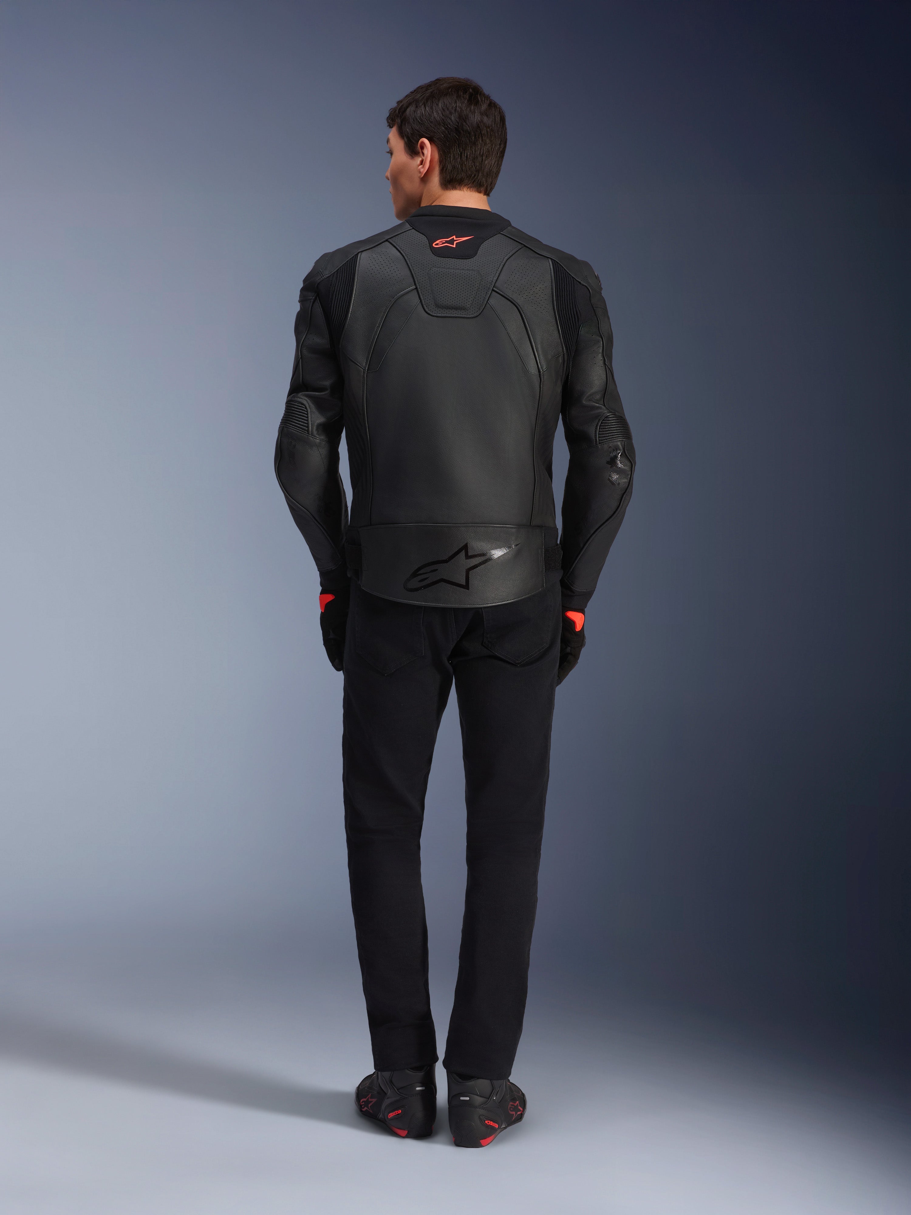 Alpinestars GP Plus R V4 Airflow Leren Jas, zwart, achteraanzicht van motorrijder in volledige uitrusting, voorzien van geperforeerd leer voor ventilatie en rode logo-accenten, gestroomlijnd sport-fit ontwerp, gecombineerd met een zwarte broek en motorlaarzen