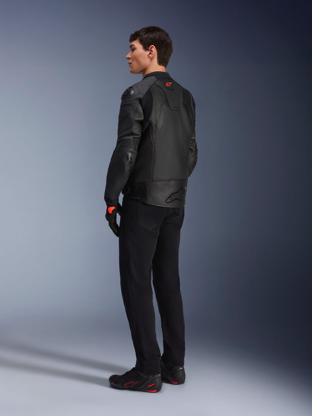 Alpinestars GP Plus R V4 Airflow leren jas, sportieve motorjas voor heren, volledig zwart, driekwart achteraanzicht op een model, voorzien van geperforeerde leren panelen, rood logo op de rug en geïntegreerde schouderbescherming, gedragen met een zwarte broek en motorschoenen
