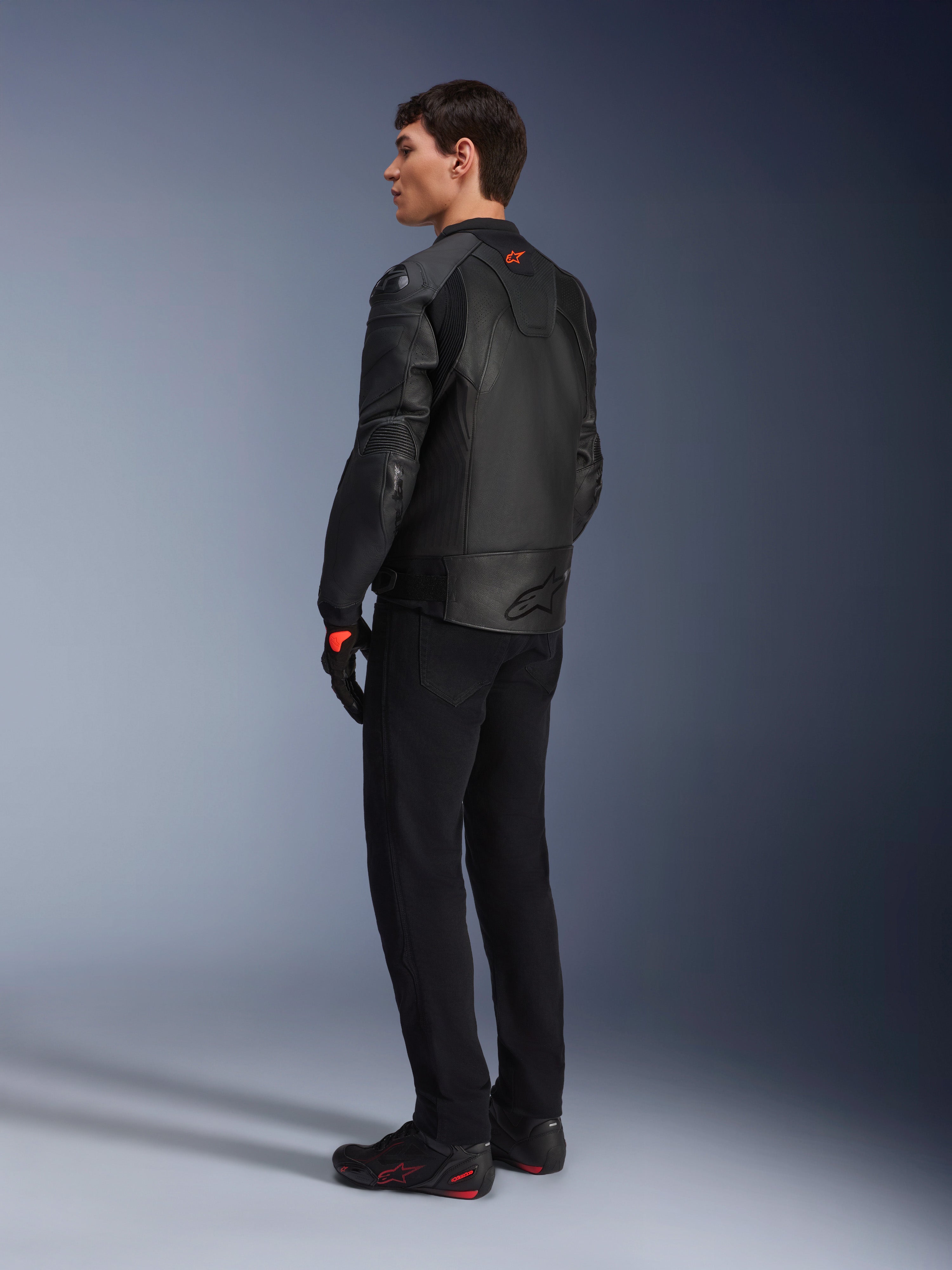 Alpinestars GP Plus R V4 Airflow leren jas, sportieve motorjas voor heren, volledig zwart, driekwart achteraanzicht op een model, voorzien van geperforeerde leren panelen, rood logo op de rug en geïntegreerde schouderbescherming, gedragen met een zwarte broek en motorschoenen