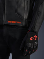 Alpinestars GP Plus R V4 Airflow leren jas, close-up van zwart geperforeerd leer met oranje grafische tekst, gecombineerd met een zwarte en oranje motorhandschoen met het Astars-logo