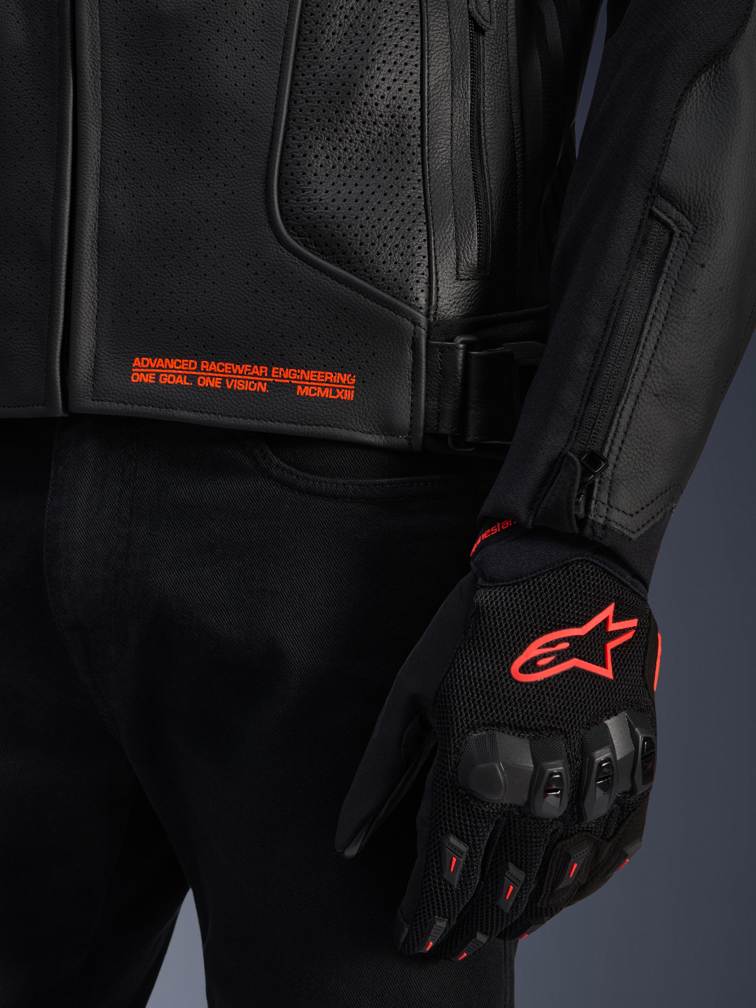 Alpinestars GP Plus R V4 Airflow leren jas, close-up van zwart geperforeerd leer met oranje grafische tekst, gecombineerd met een zwarte en oranje motorhandschoen met het Astars-logo