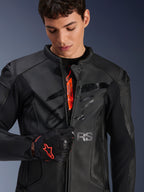 Alpinestars GP Plus R V4 Airflow leren jas in zwart, mannelijk model ritst de voorzijde gedeeltelijk open om de oranje branding aan de binnenkant te tonen, met geperforeerde leren panelen en een glanzend logo op de borst, gecombineerd met een zwart-rode motorhandschoen