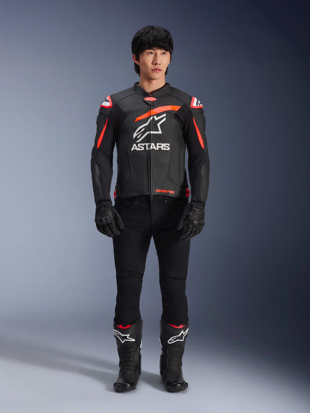 Mannelijk model draagt Alpinestars GP Plus R V4 Airflow lederen jas, zwart met rood fluo en witte accenten, groot wit Astars-logo op de borst, gecombineerd met zwarte technische broek, handschoenen en laarzen, performance motoruitrusting in een studio-omgeving