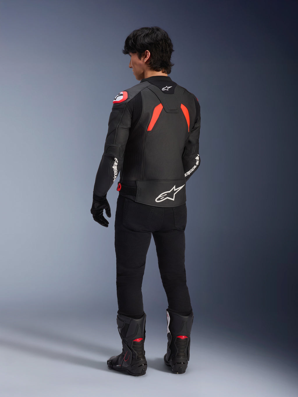 Achteraanzicht van een model in de Alpinestars GP Plus R V4 Airflow lederen jas, zwart met Red Fluo en witte accenten, gedragen met een zwarte broek en zwarte motorlaarzen, met opvallende witte logo's op de rug en mouwen, sportieve lederen constructie
