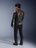 Achteraanzicht van een model in de Alpinestars GP Plus R V4 Airflow lederen jas, zwart met Red Fluo en witte accenten, gedragen met een zwarte broek en zwarte motorlaarzen, met opvallende witte logo's op de rug en mouwen, sportieve lederen constructie