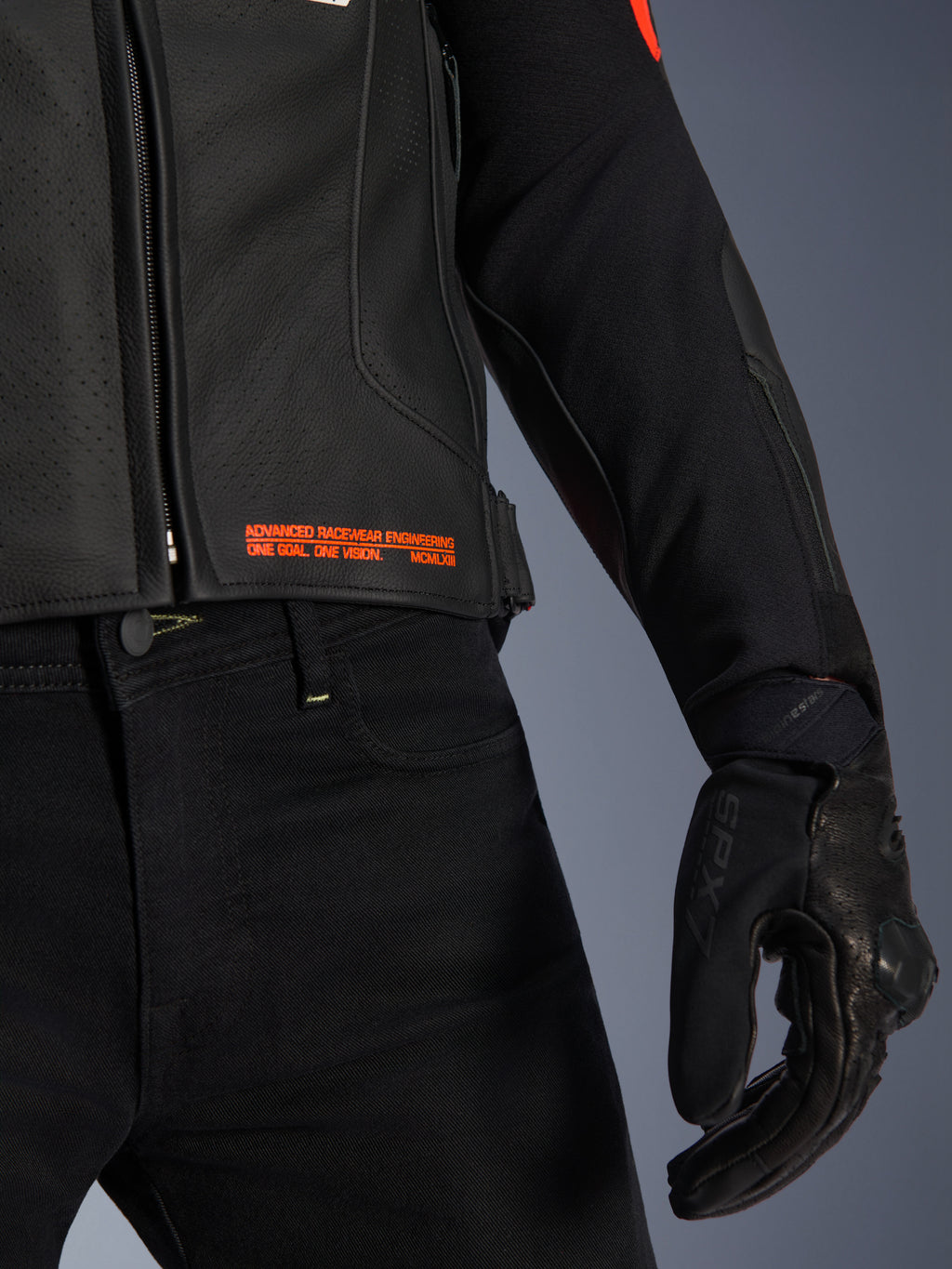 Alpinestars GP Plus R V4 Airflow leren jas, close-up van de taille en mouw, zwart geperforeerd leer met fluo rode tekst 'Advanced Racewear Engineering', gecombineerd met een zwarte motorbroek en SPX 7 motorhandschoen