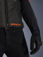 Alpinestars GP Plus R V4 Airflow leren jas, close-up van de taille en mouw, zwart geperforeerd leer met fluo rode tekst 'Advanced Racewear Engineering', gecombineerd met een zwarte motorbroek en SPX 7 motorhandschoen