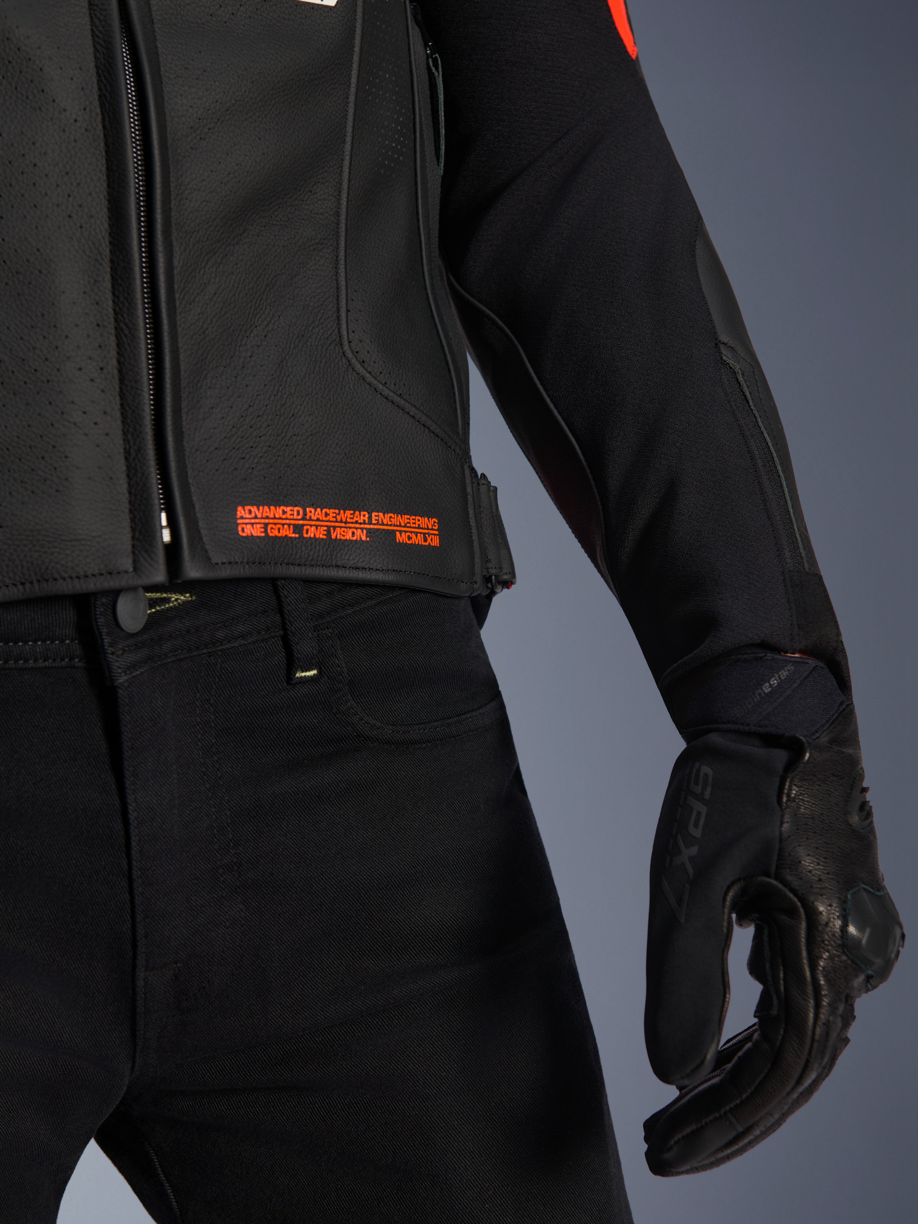 Alpinestars GP Plus R V4 Airflow leren jas, close-up van de taille en mouw, zwart geperforeerd leer met fluo rode tekst 'Advanced Racewear Engineering', gecombineerd met een zwarte motorbroek en SPX 7 motorhandschoen