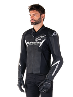 Alpinestars Faster V3 leren jas, sportieve motorjas, zwart en wit, prominent wit Alpinestars-logo en 'Racing Ahead'-tekst op de borst, sterlogo's op de schouders, hoogwaardige leren constructie met stretchpanelen, gedragen door een model met zwarte broek en handschoenen