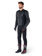 Man die Alpinestars Faster V3 Airflow leren jas in Black Black draagt, gecombineerd met een zwarte broek, zwart-rode motorhandschoenen en zwarte racelaarzen met rode en witte accenten, volledige sportieve motoruitrusting