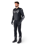 Model draagt Alpinestars Faster V3 Airflow Leren Jas, zwart en wit, voorzien van geperforeerd leer en witte Alpinestars-logo's, gecombineerd met een zwarte motorbroek, handschoenen en laarzen, sportieve motoruitrusting