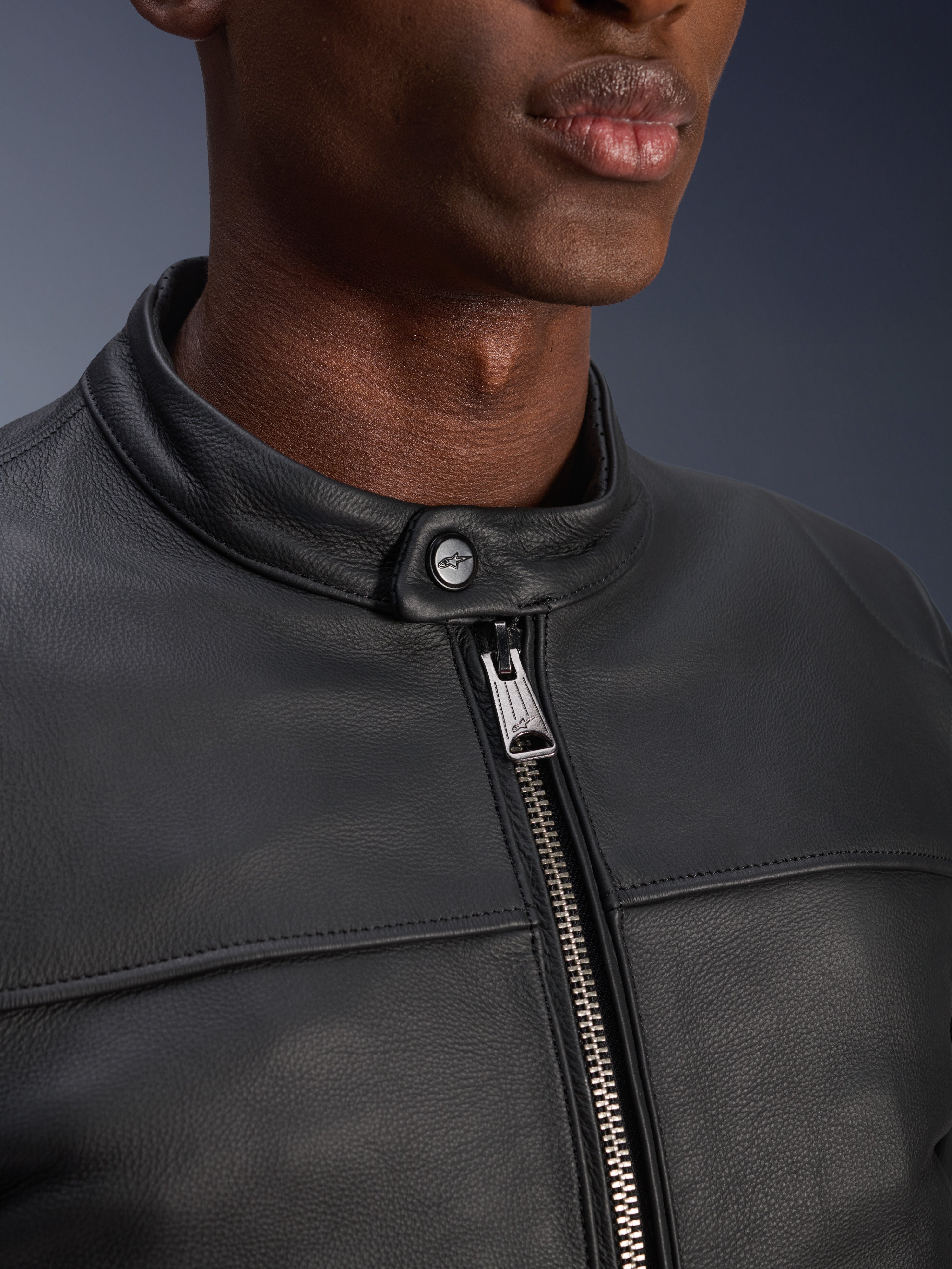 Close-up van de Alpinestars Blacktrack Leren Jas, met de metallic gunmetal rits en knoopsluiting bij de nek met Alpinestars-logo, premium zwarte rundleertextuur, urbane motorkleding